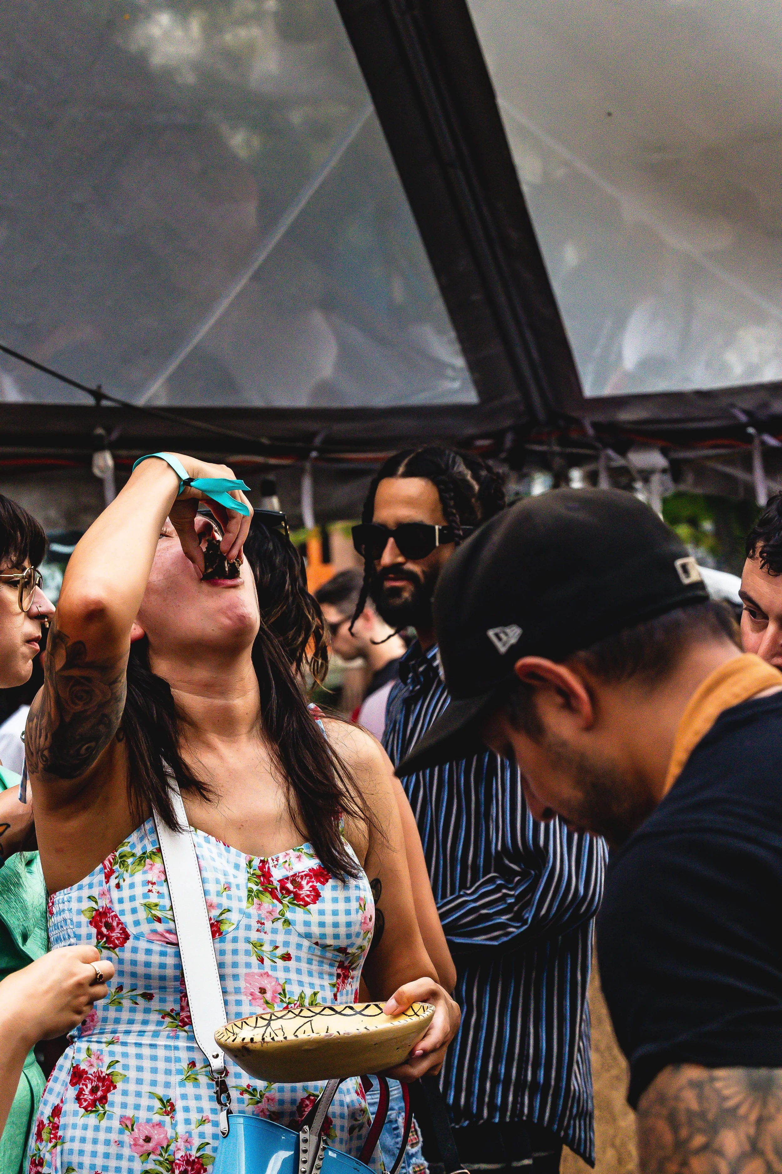 A woman in a floral dress takes a shot of a drink at an outdoor festival, with several people around her, including a man with sunglasses and a woman with glasses, under a canopy.