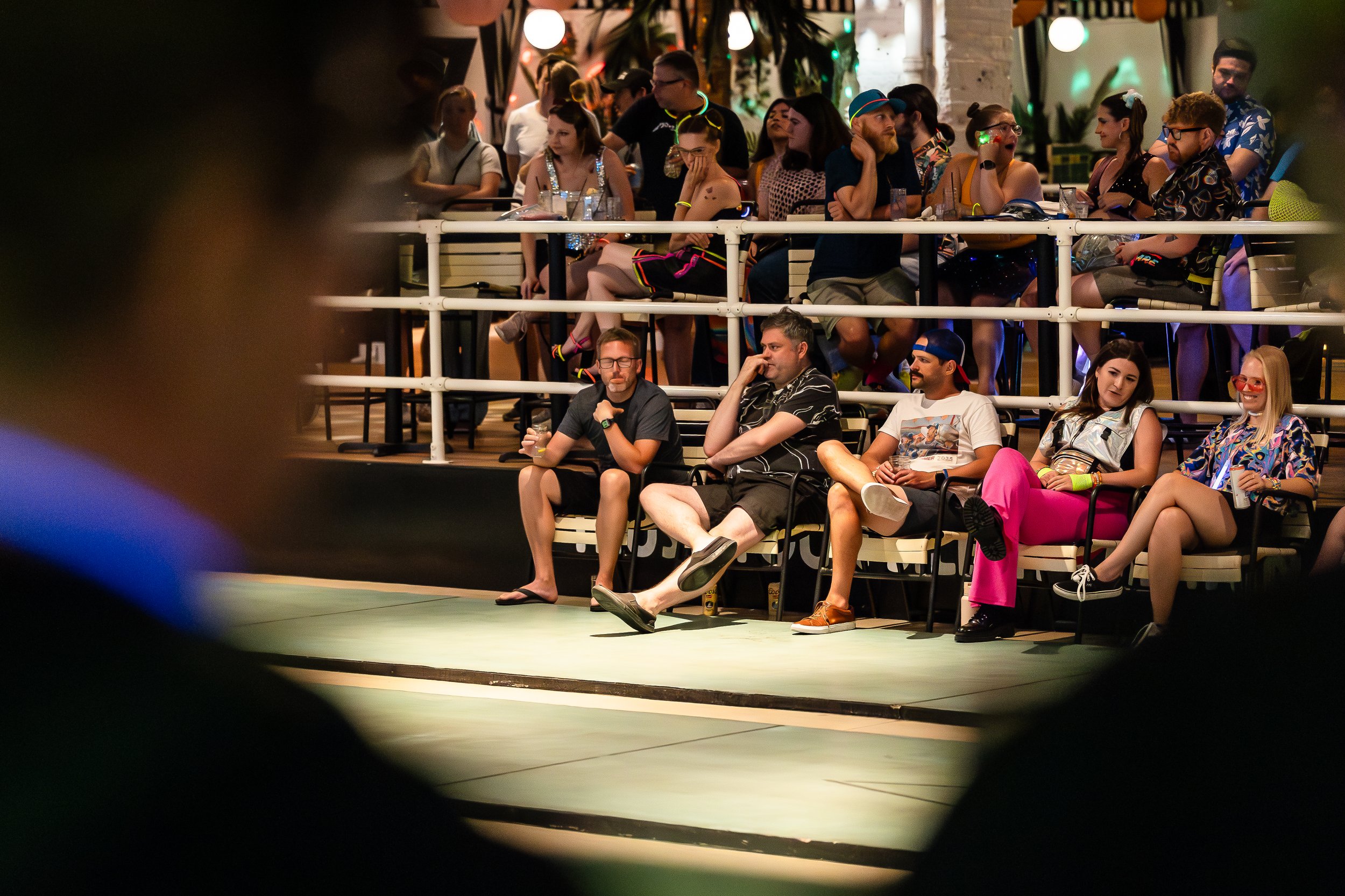 Audience members sitting and standing in an indoor venue, dressed in colorful, casual, and fashionable clothing, with some wearing glow-in-the-dark accessories, during a lively event or performance.