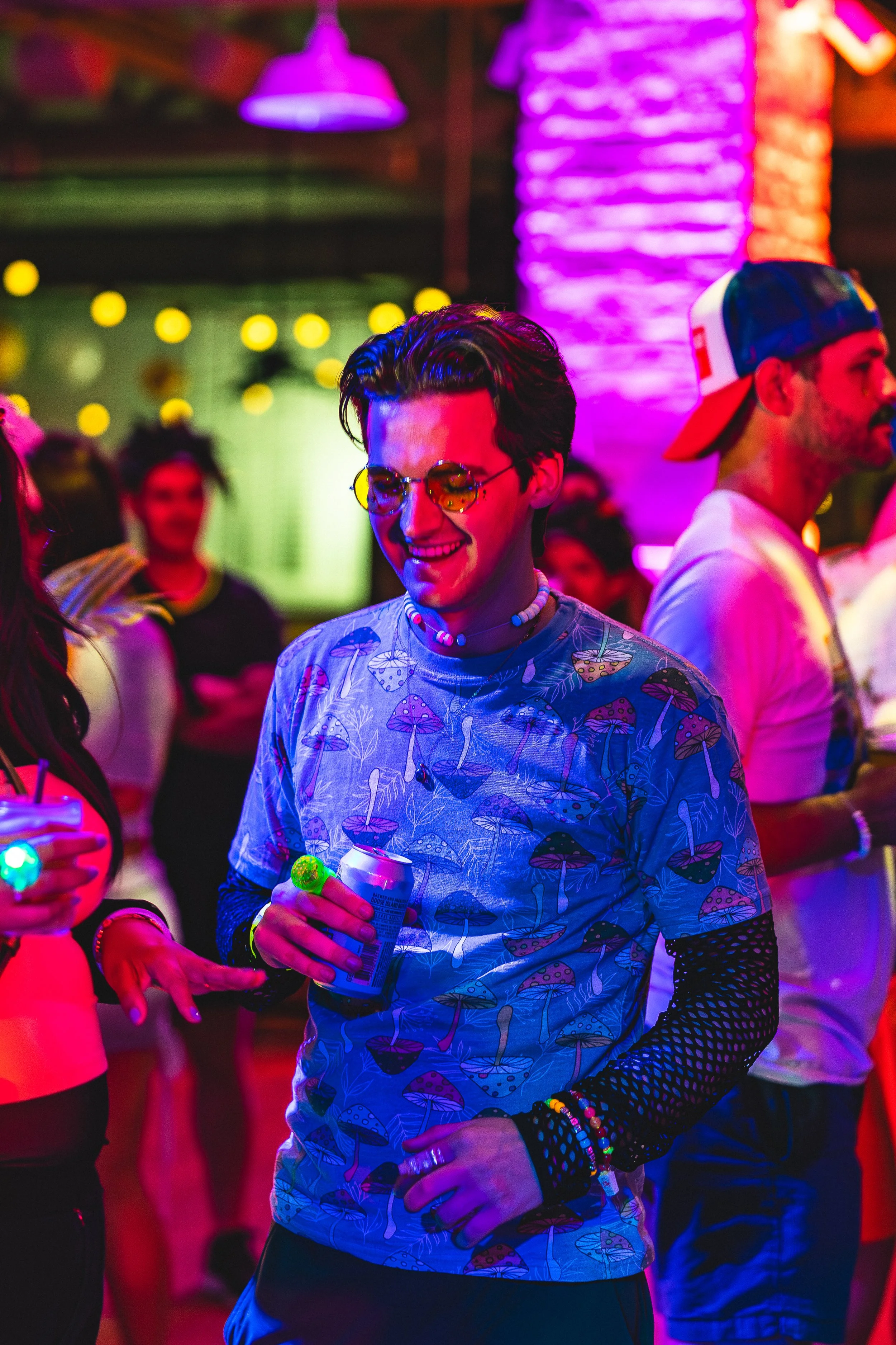 Person at a party wearing sunglasses, a mushroom-printed shirt, and holding a can of beverage, surrounded by colorful neon lighting