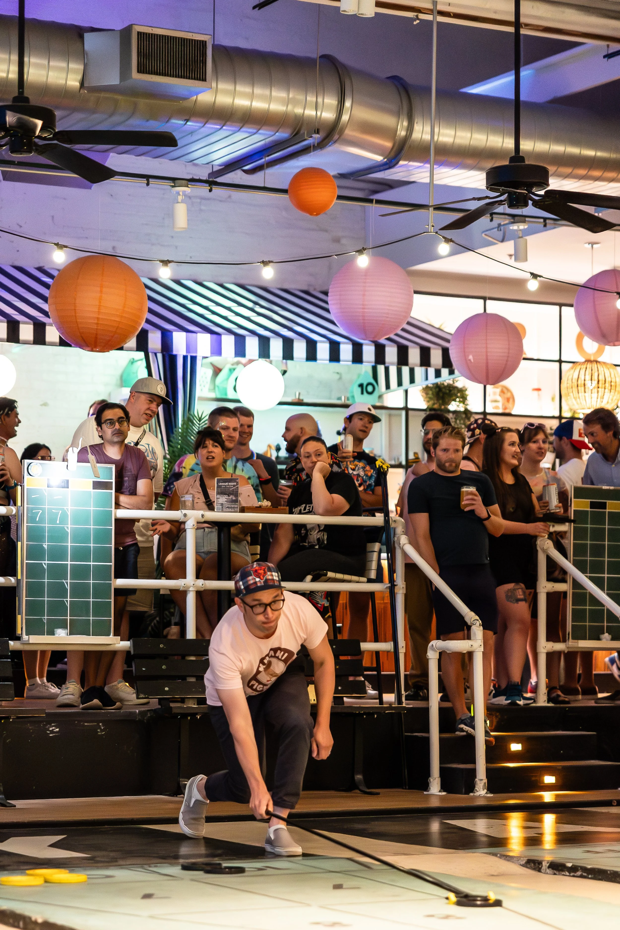 A group of people watching a shuffleboard game at an indoor venue decorated with pink, orange, and white paper lanterns, and string lights. One person is crouching, holding a shuffleboard cue, preparing to slide a puck on the court floor.