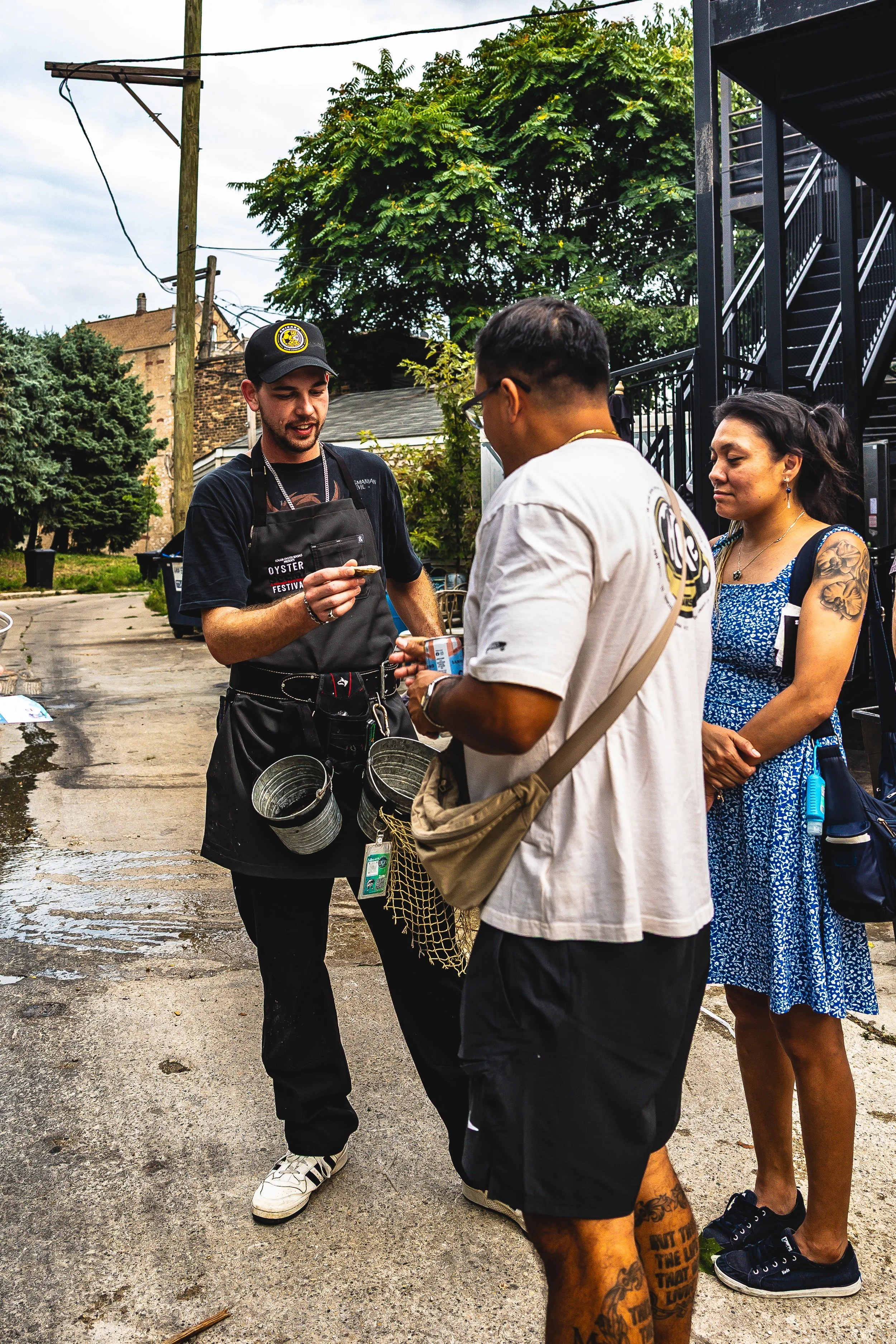 Three people, two men and one woman, standing on a wet street outdoors, engaged in conversation. The man on the left is wearing a black cap and apron, holding a small object, possibly an oyster. The man in the middle wears glasses and a white t-shirt