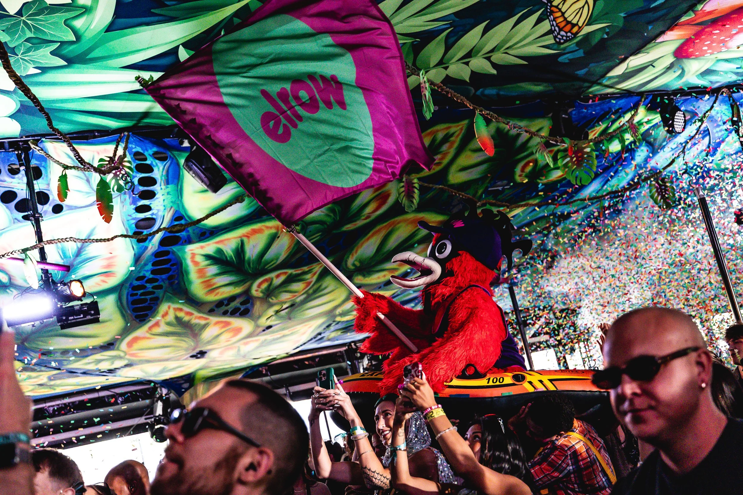 Crowd of people at a festival with a person in a crow costume holding a pink and green flag on a decorated float covered with colorful, psychedelic patterns and confetti.