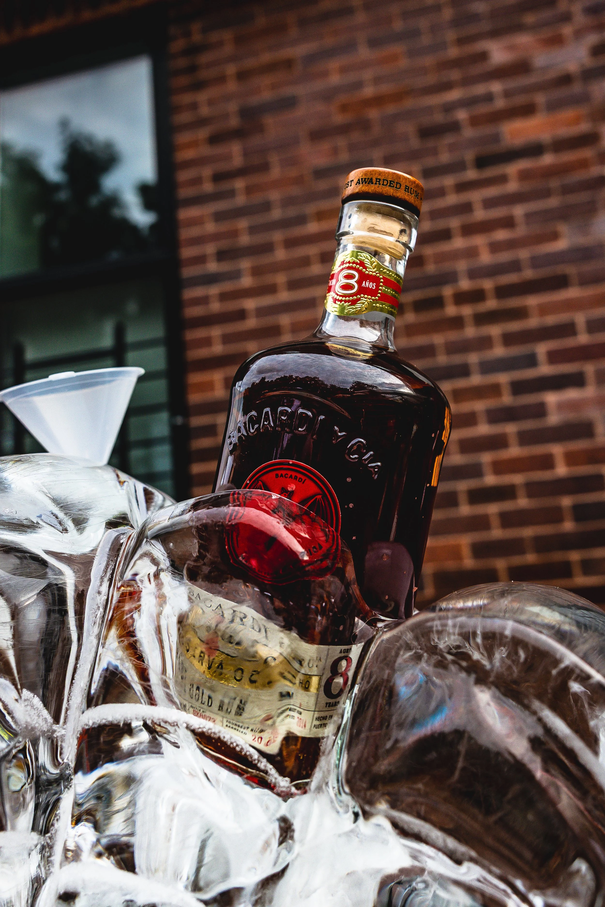 A bottle of Bacardi rum partially submerged in ice in an outdoor setting with a red brick building in the background.