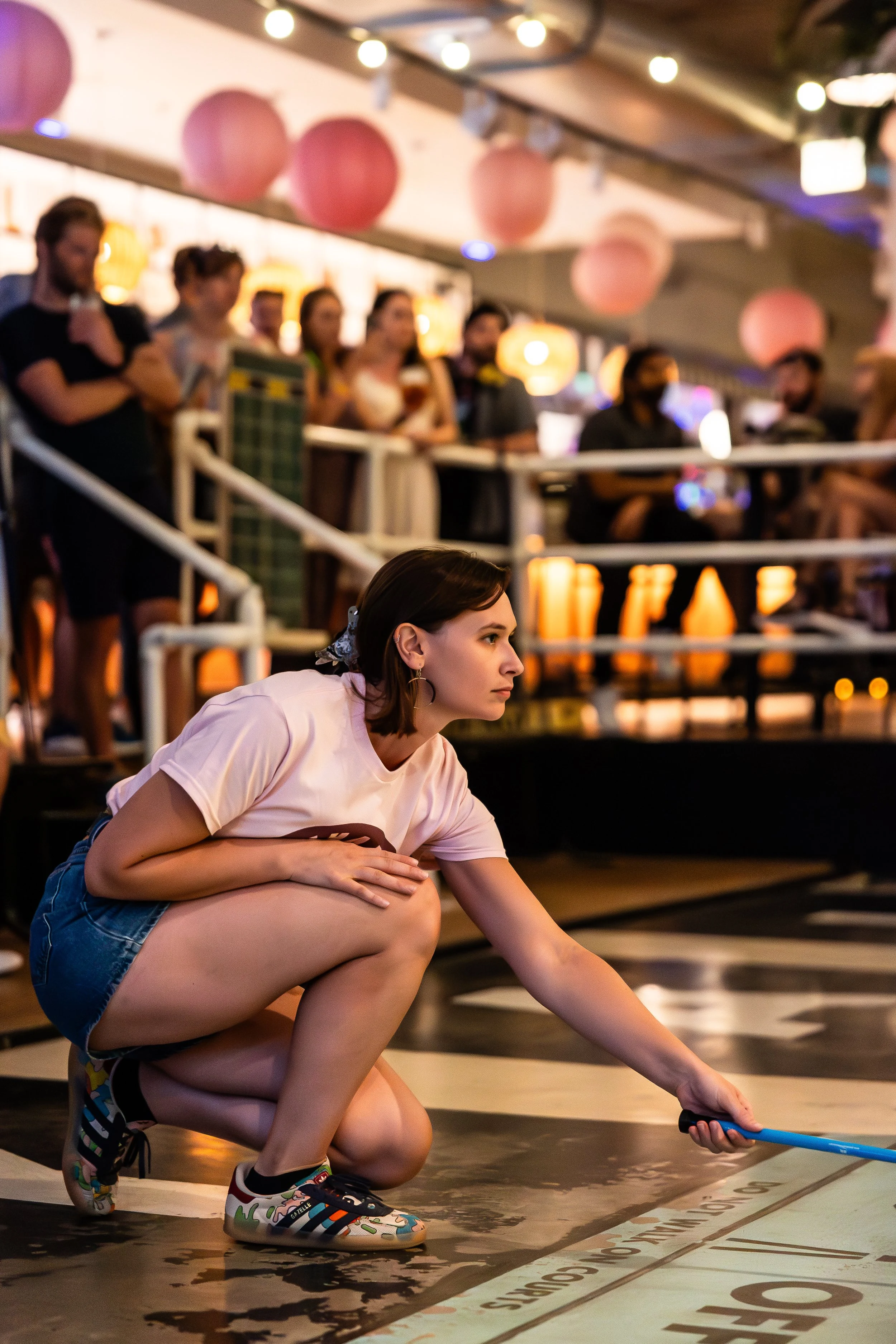 A woman crouching down and cleaning the floor of a bowling alley with a mop, while a group of people watch from the background.