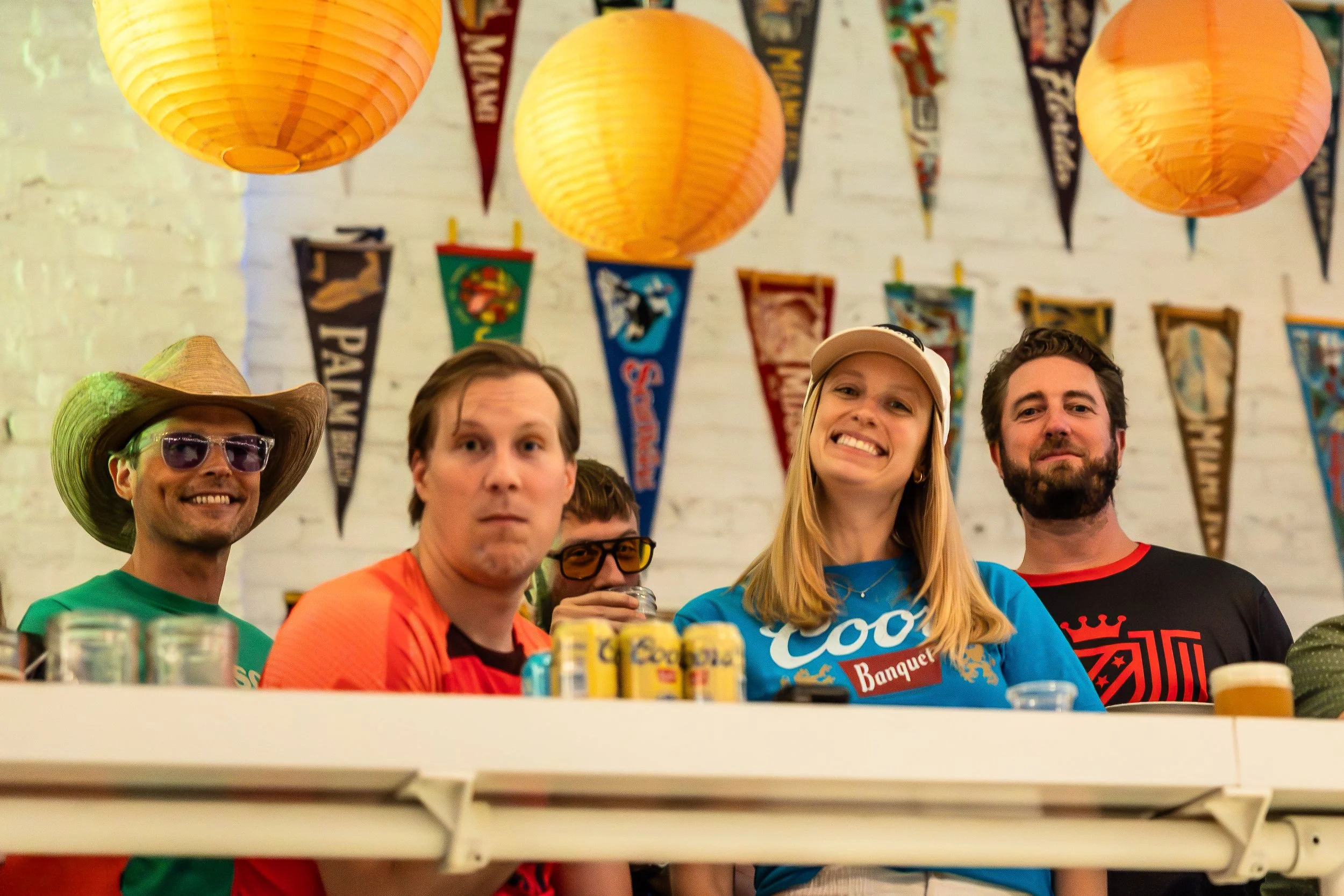 A group of five friends gathered around a table, smiling, with colorful lanterns and sports flags hanging on a white brick wall in the background, celebrating together.