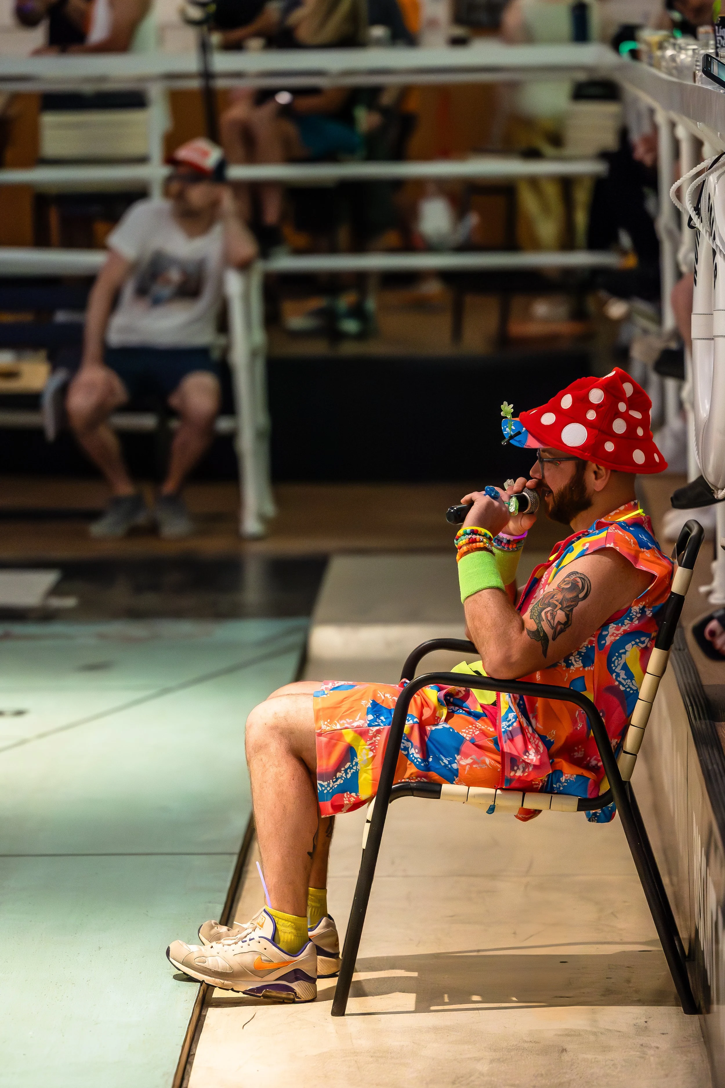 A person dressed in a colorful, tropical-themed outfit with a red polka-dot hat, sitting on a chair in a humorous pose, talking on a walkie-talkie inside a bowling alley.