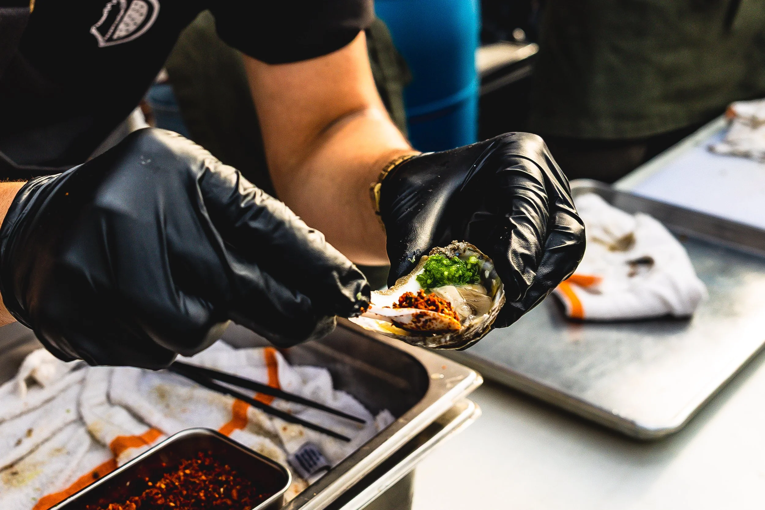 A person wearing black gloves preparing a seafood oyster with toppings at a food stand