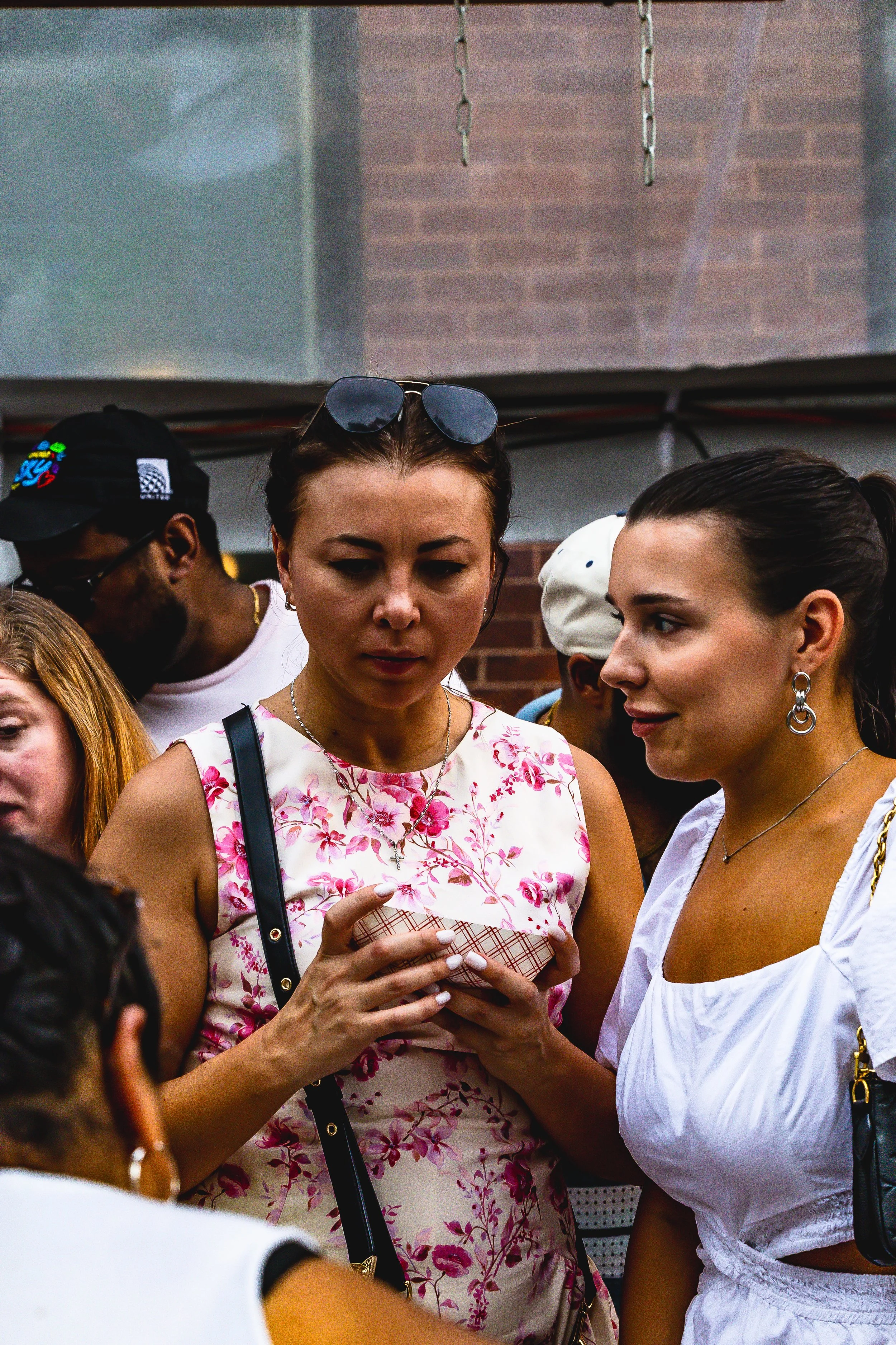 Two women are looking at a phone closely in a crowd, with a brick wall and ceiling in the background.