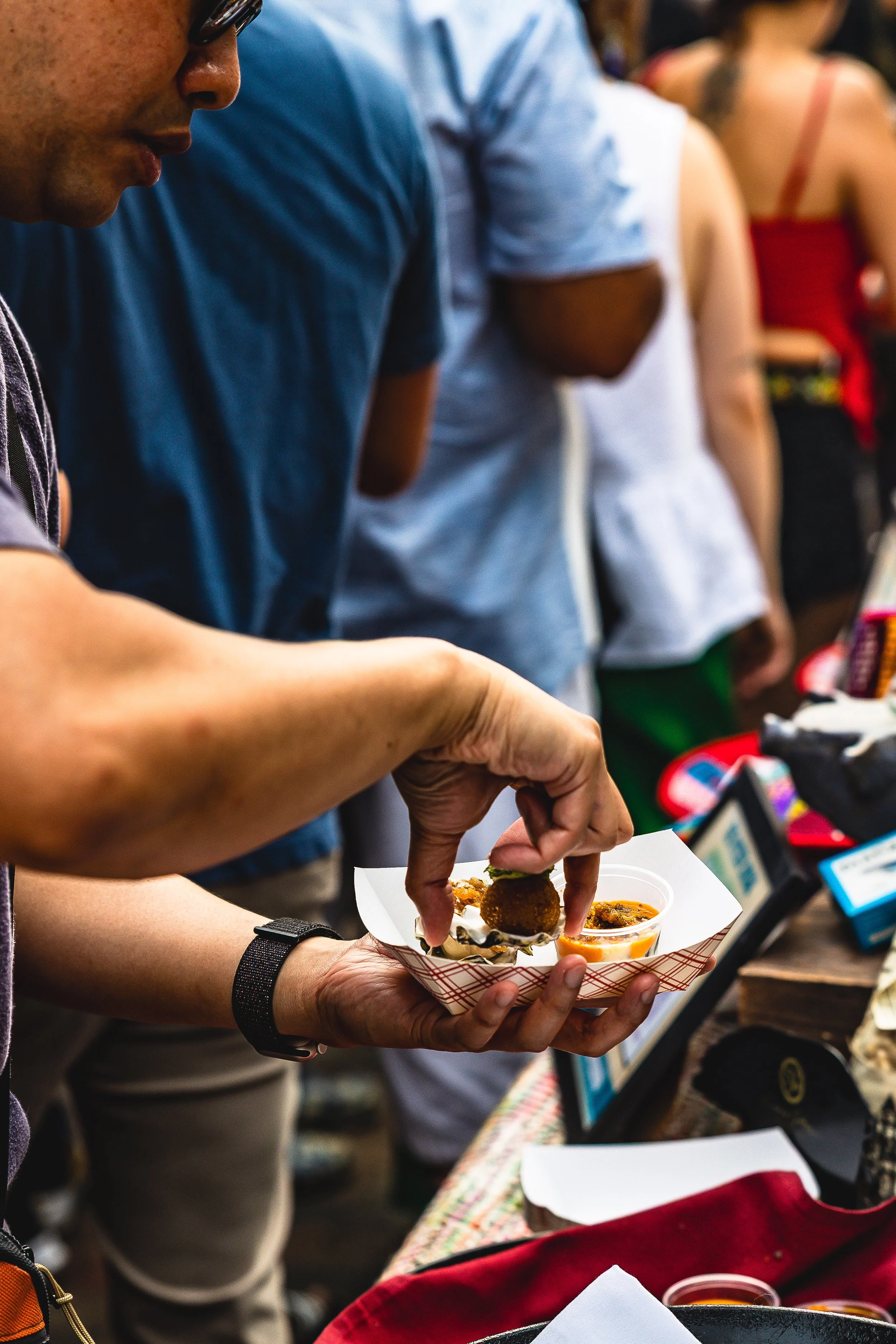 A person holding a small food sample in a white paper tray at a food event, with other people in line behind them.