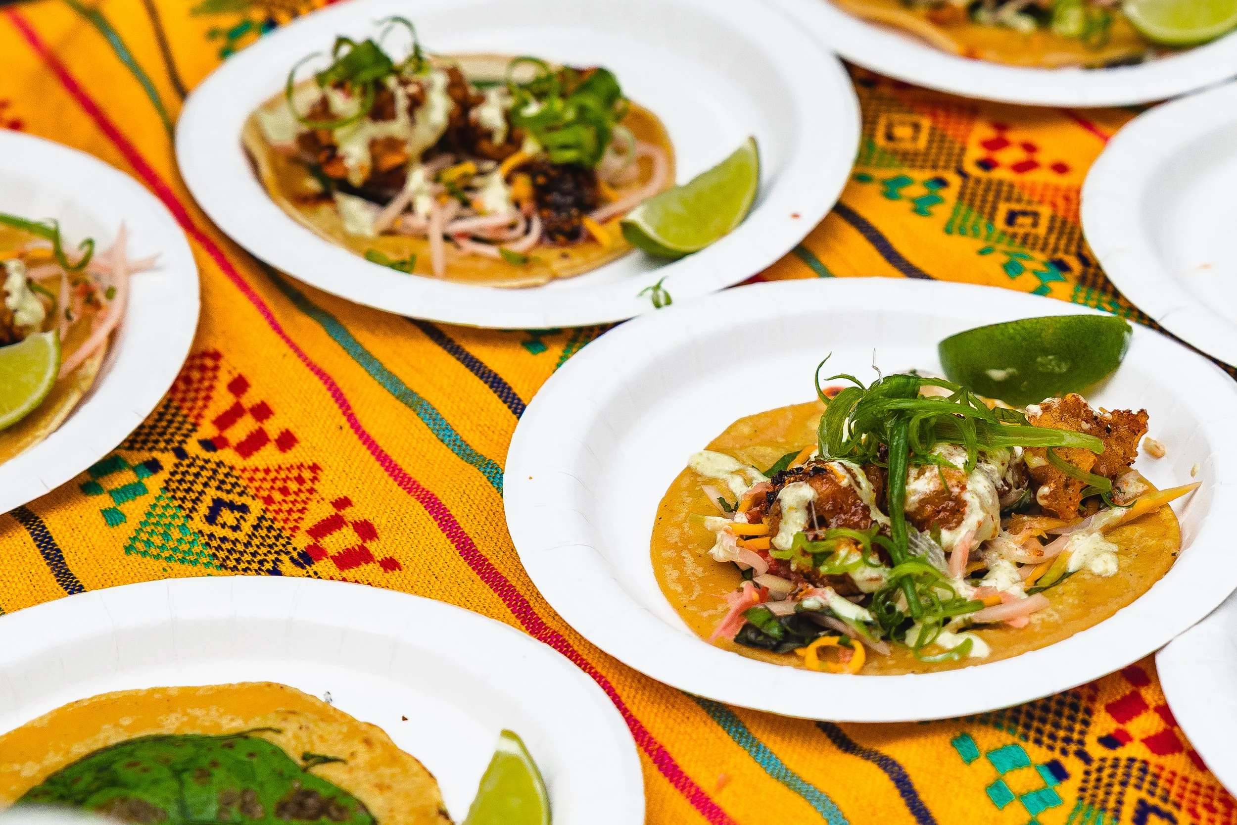 Several plates of tacos on a colorful tablecloth, each topped with meats, vegetables, and garnished with lime wedges.