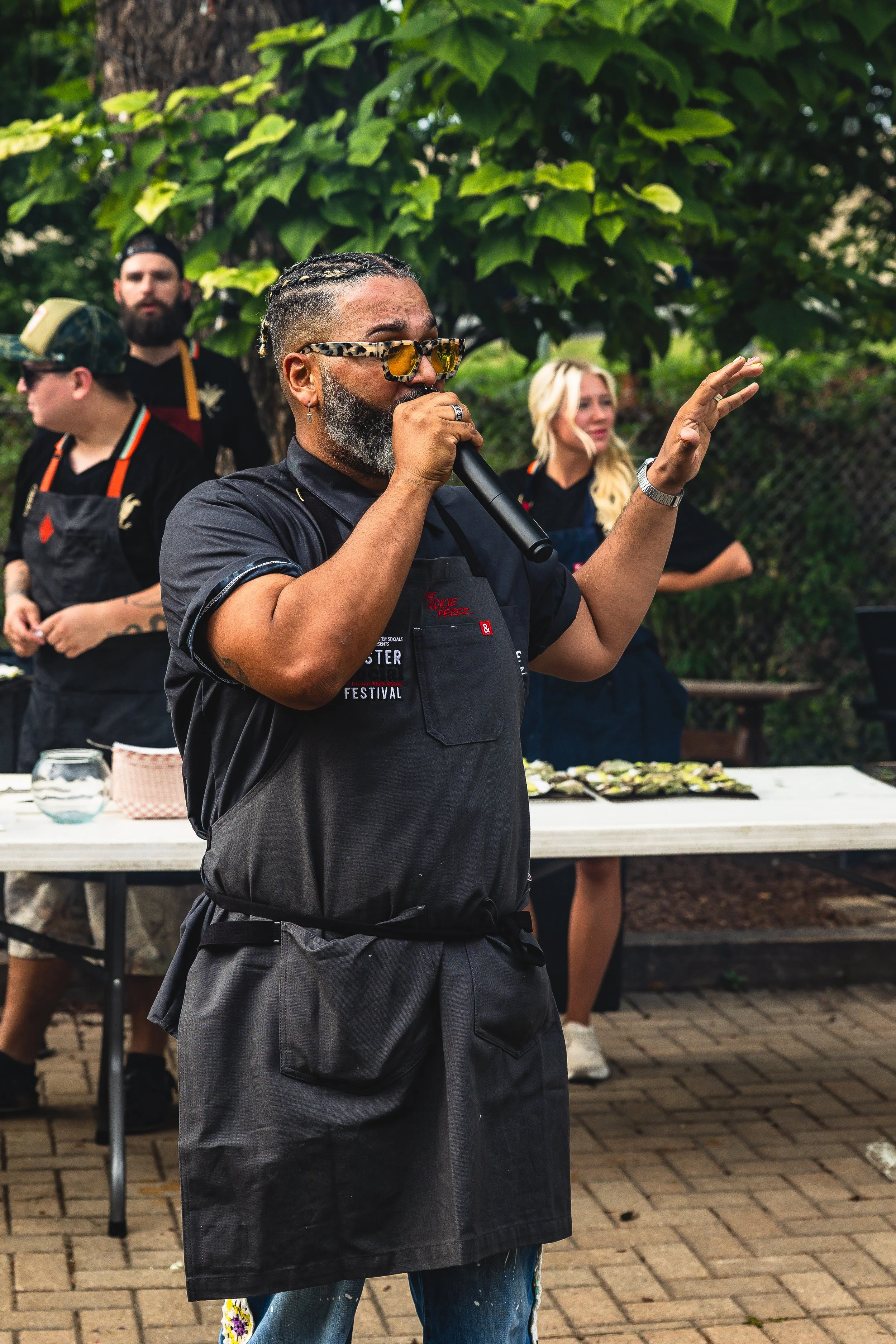 A man with gray beard, glasses, and braided hair speaking into a microphone at an outdoor event, surrounded by a group of people, some wearing aprons, with lush green foliage in the background.