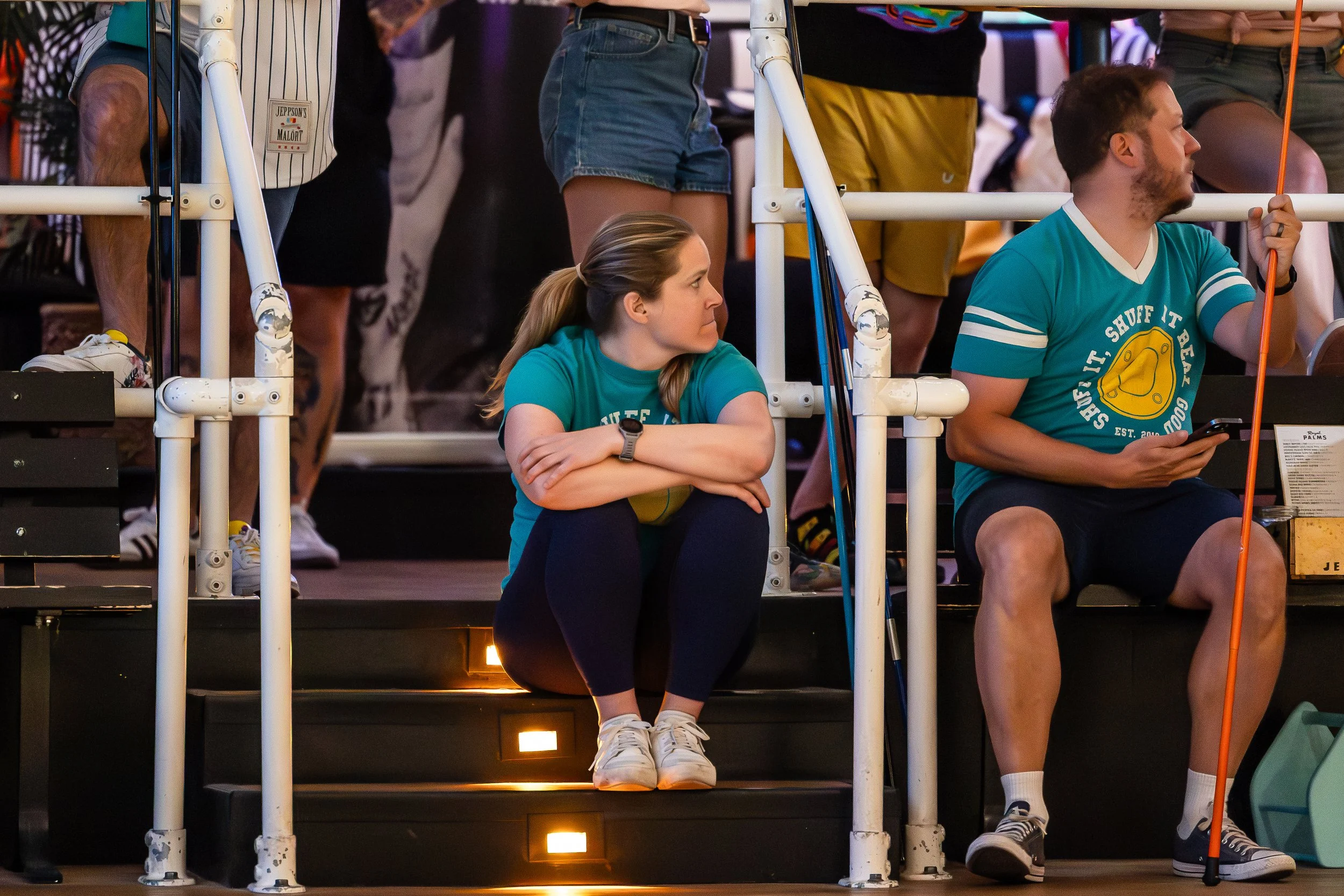 A woman sitting on the stairs at an indoor event, wearing a turquoise T-shirt, dark leggings, and white sneakers, with arms crossed and looking to her left. Next to her, a man in a turquoise T-shirt, shorts, and sneakers is sitting on the stairs, hol