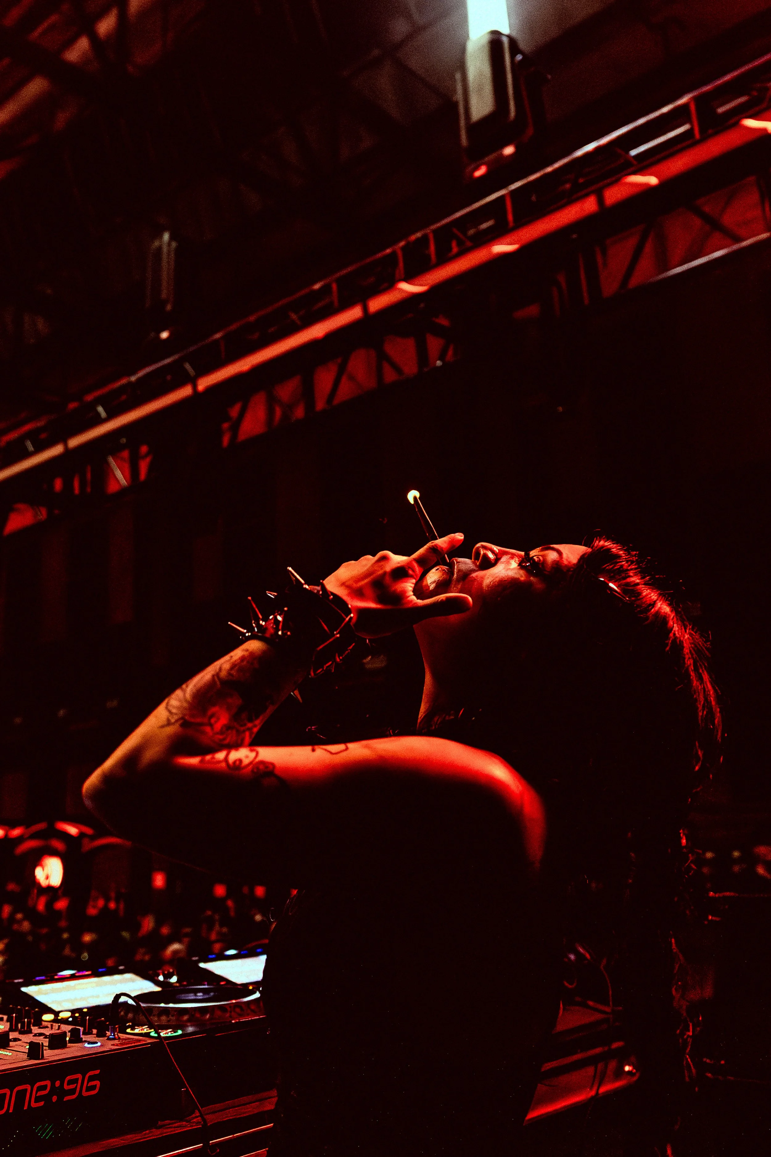 A female DJ performing in a dark, red-lit nightclub, adjusting her headphones with one hand while the DJ mixer and turntables are visible in front of her.