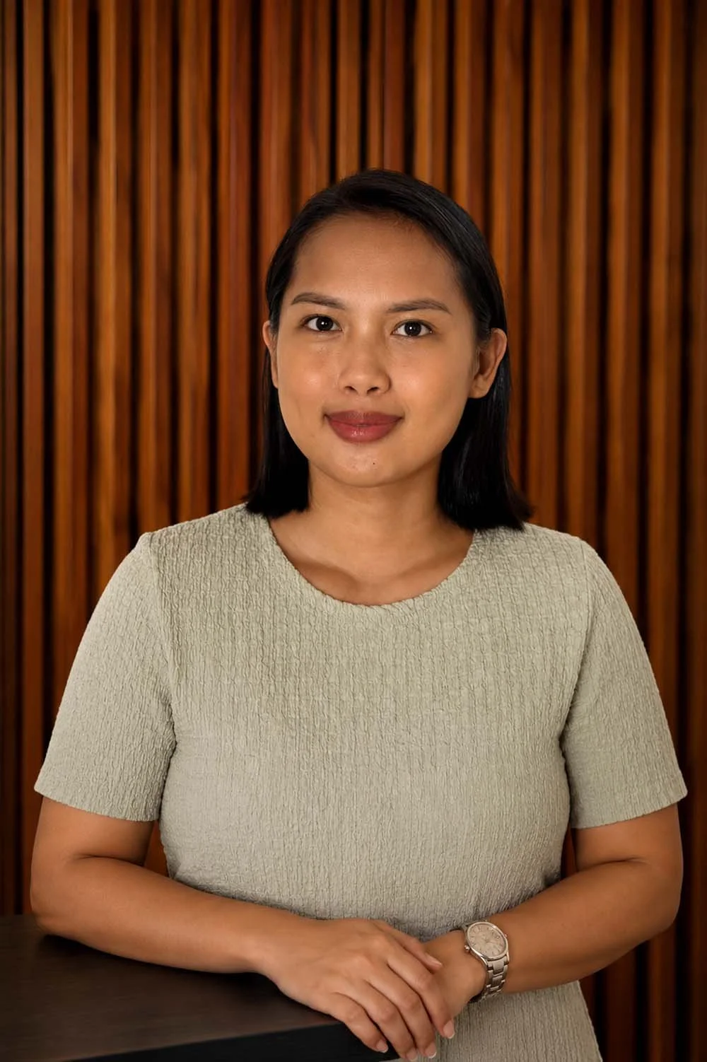 A person with short black hair wearing a textured light green top is sitting indoors with a neutral background. In the foreground, there are yellow flowers.