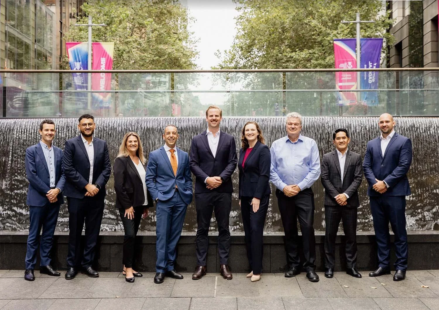 A group of 9 diverse business professionals standing side by side in front of a Sydney fountain, with trees and colourful banners in the background. This is the team at Advisers Services Hub.
