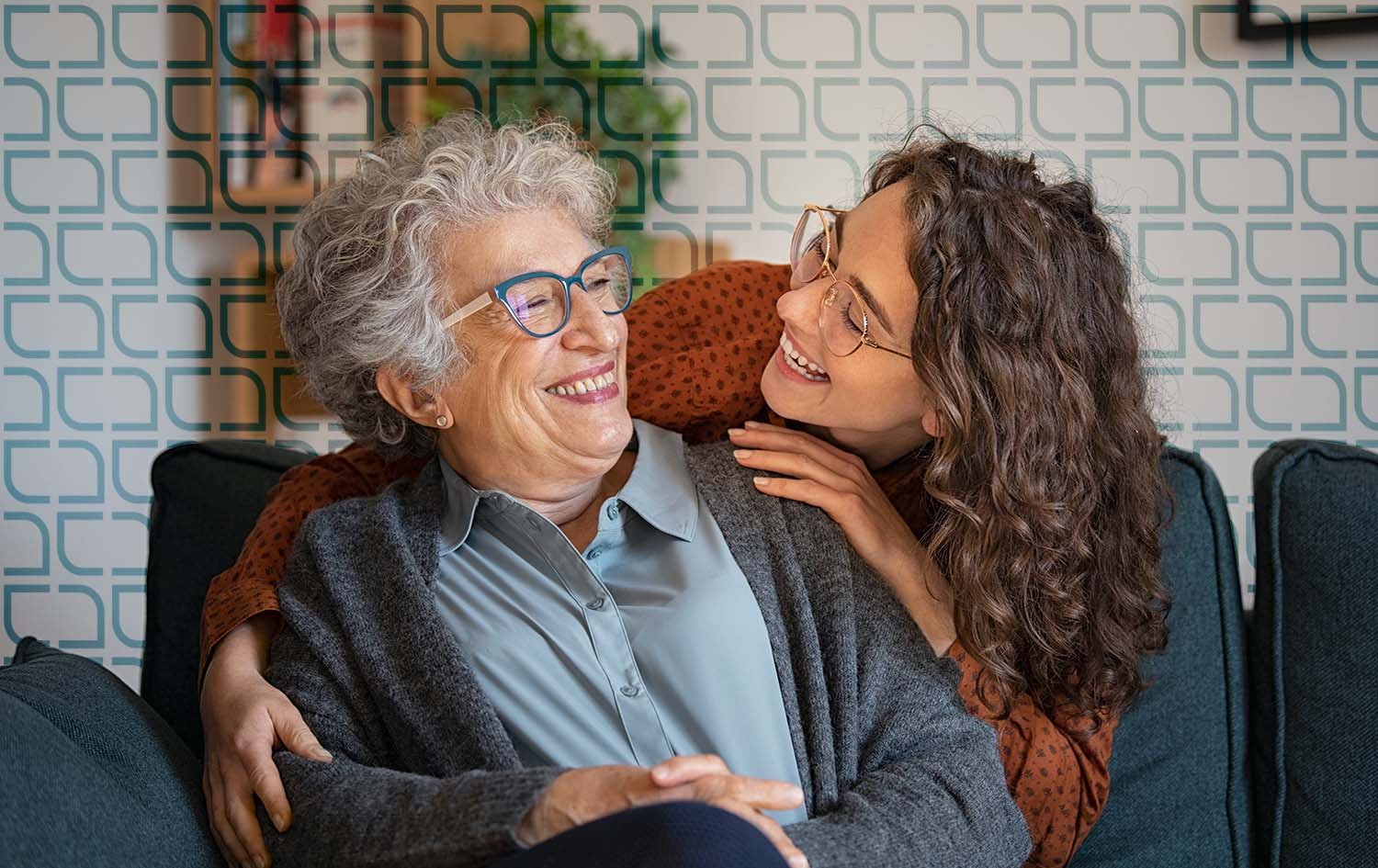 An elderly woman with gray hair and a younger woman with curly brown hair and glasses, together on a couch, smiling and sharing a moment of affection while discussing aged care living.