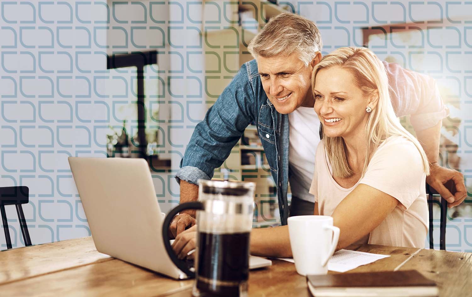 man and a woman, look at a laptop screen together in a bright, modern kitchen while on a virtual call with their financial advisor with a mug, a French press coffee maker, and a notebook nearby. They are smiling and enjoying their time.