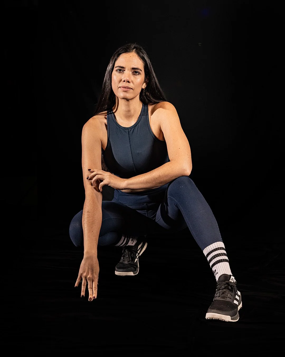 Mujer en postura de ejercicio, agachada con un brazo extendido hacia abajo y la otra cruzada sobre su rodilla, viste ropa deportiva negra y medias blancas con rayas negras, fondo negro.