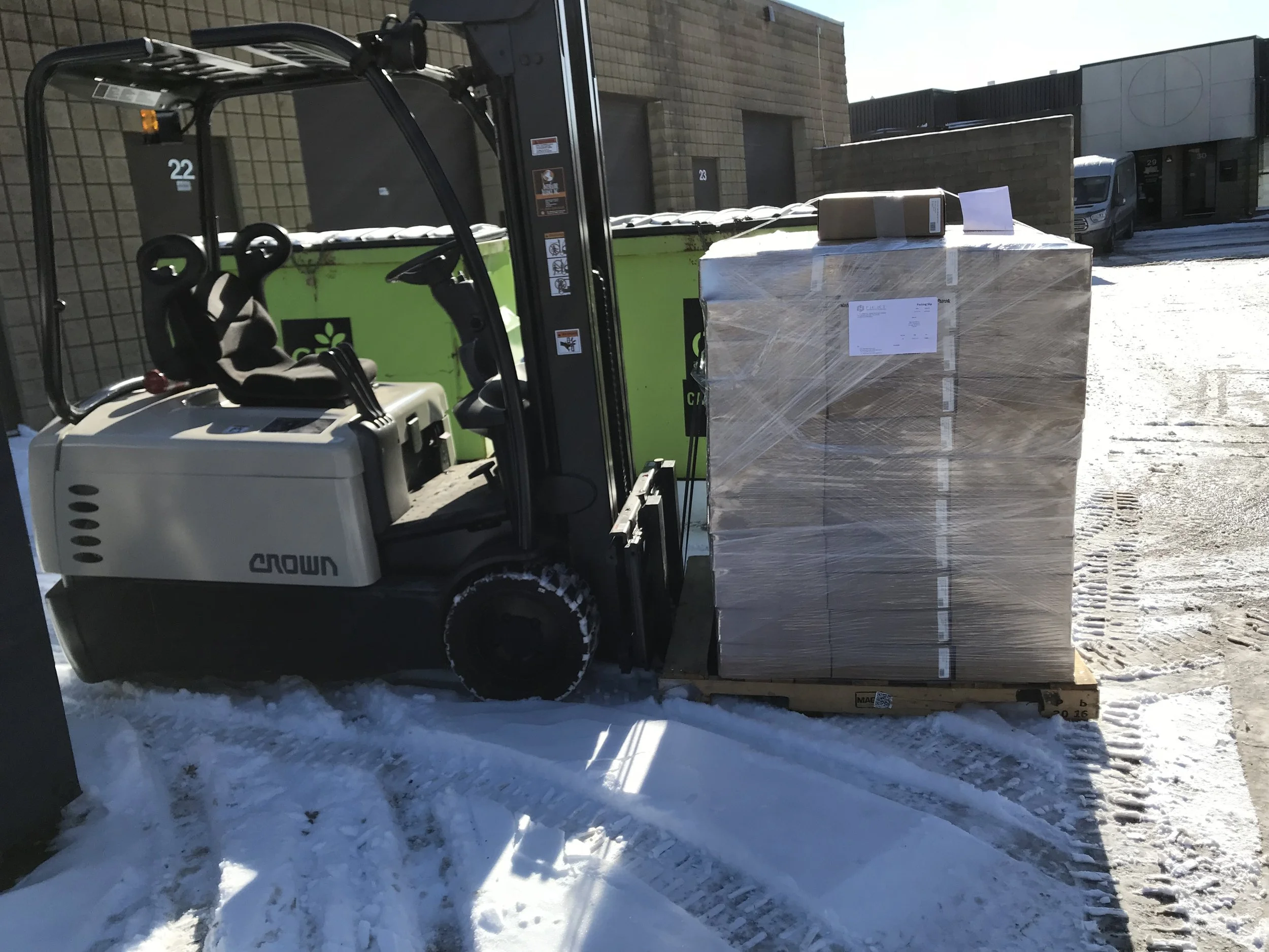 Forklift moving a wrapped pallet of boxes on a snowy ground outside a warehouse.