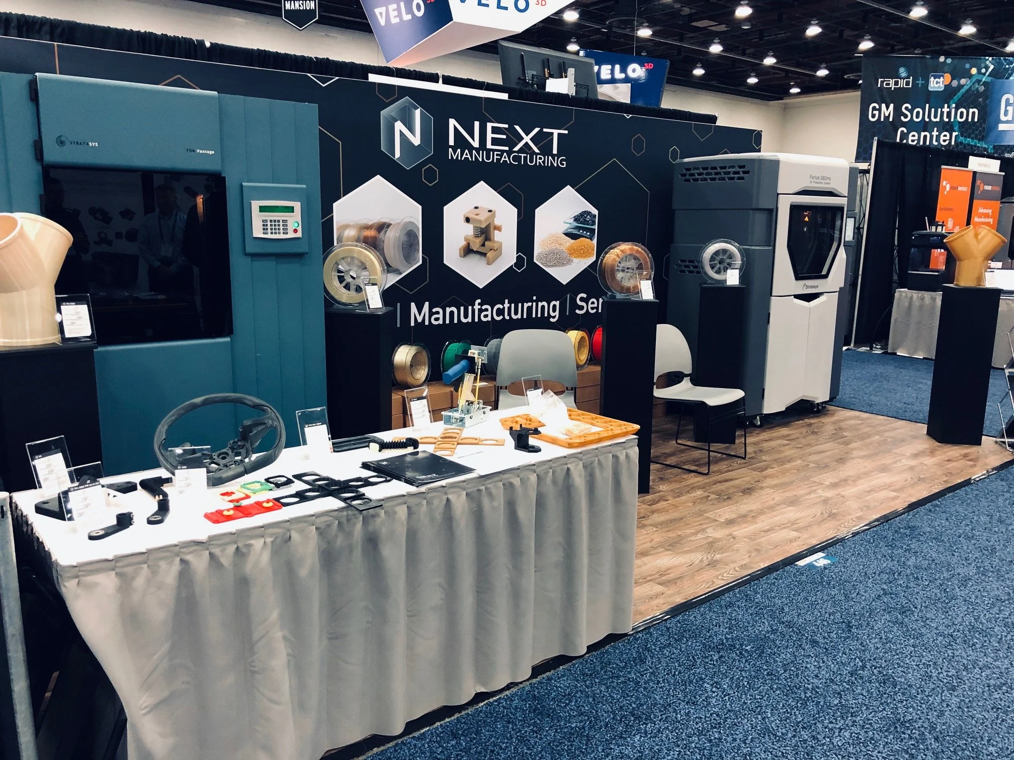 NEXT 3DP booth at RAPID + TCT 2019 in Michigan, featuring Stratasys Fortus 450mc and Fortus 400mc systems printing Stratasys-compatible FDM materials, including an ABS-M30® alternative and a custom TPU, with functional production parts on display.