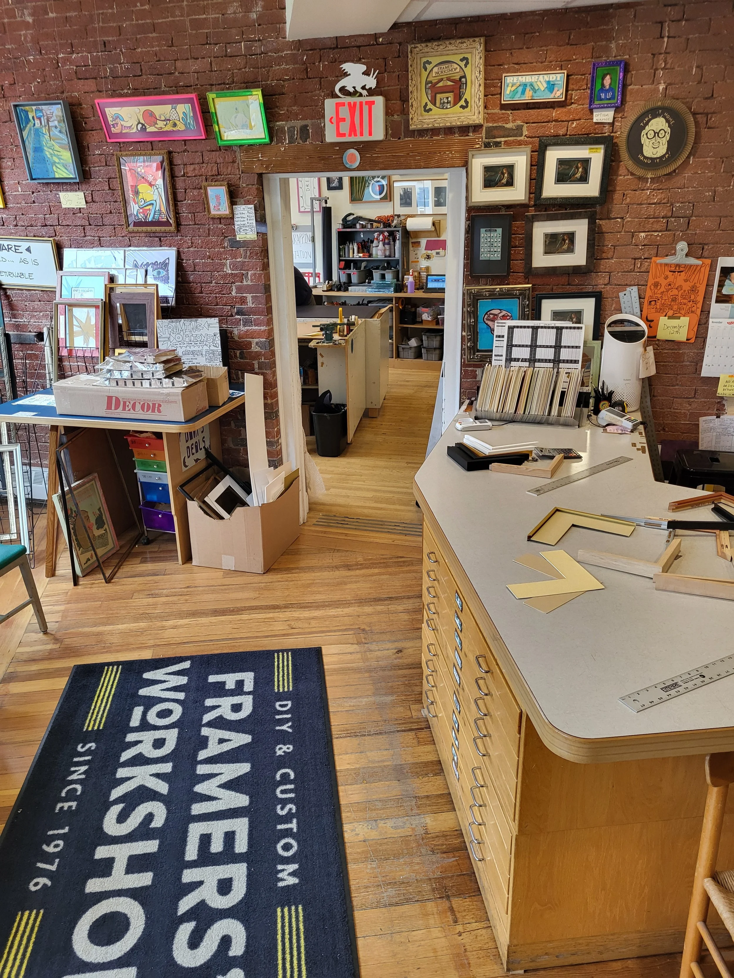 Interior of a framing shop with a brick wall, framed artwork, a black and yellow doormat reading 'Framer's Workshop, Since 1916,' a white countertop with supplies, and an open doorway leading to a back room with shelves and tools.