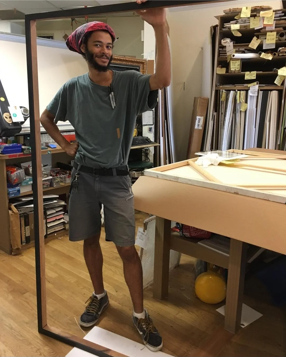 A man in a gray t-shirt, gray shorts, and sneakers looks into a mirror, taking a selfie. He is wearing a red headscarf and is smiling. The background shows a workshop with shelves filled with rolls of paper or fabric and sticky notes, and a table wit