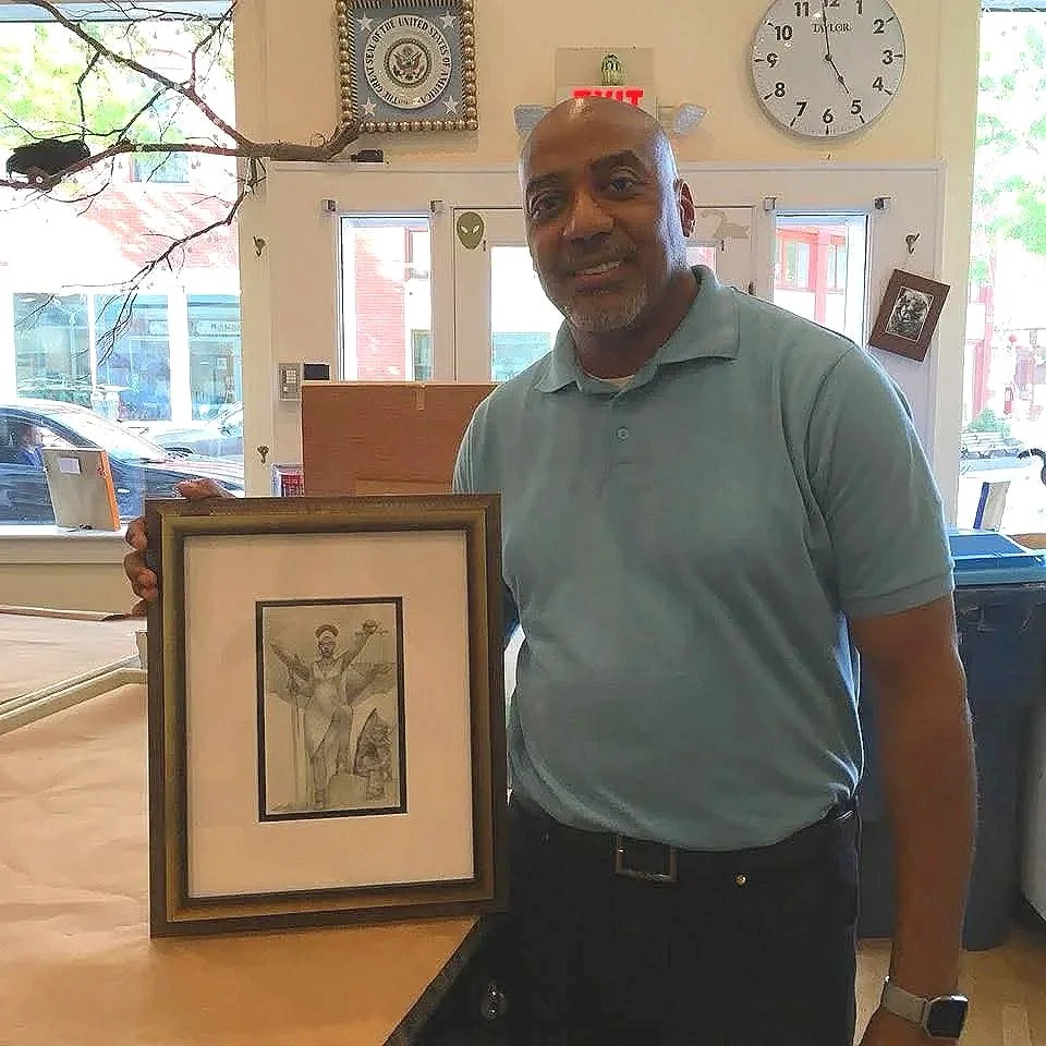 A man with a balding head and gray beard wearing a light blue polo shirt and black pants, smiling and holding a framed black-and-white drawing of a person with wings, inside a store with large windows and a clock on the wall.