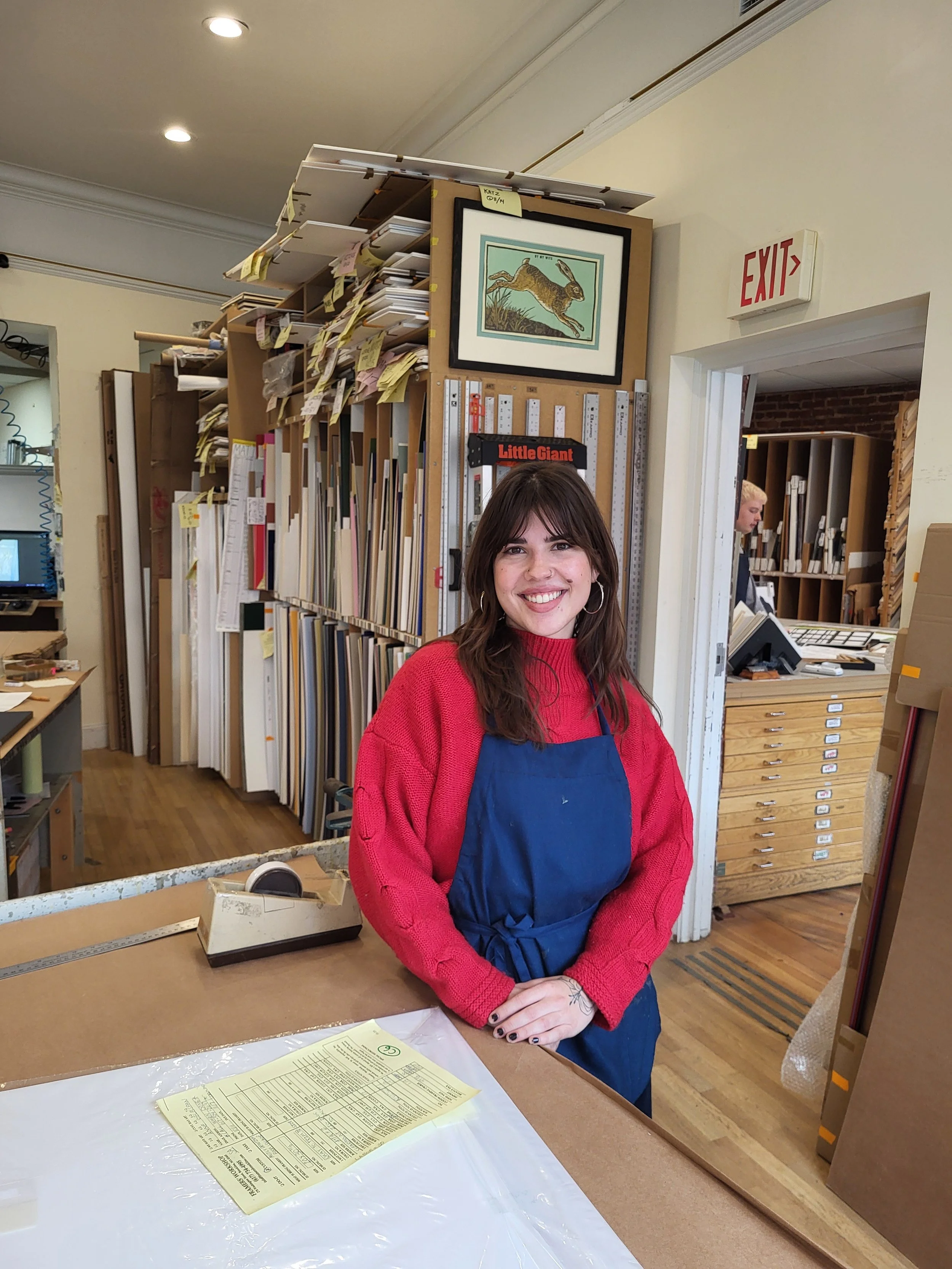 A smiling woman in a red sweater and blue apron standing behind a work counter in an art or framing shop, with shelves of artwork and framing supplies in the background.
