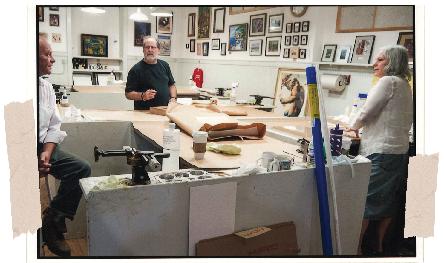 Three people in an art studio or gallery, engaged in conversation, with artwork and framed pictures on the walls, a table with art supplies, and an easel.