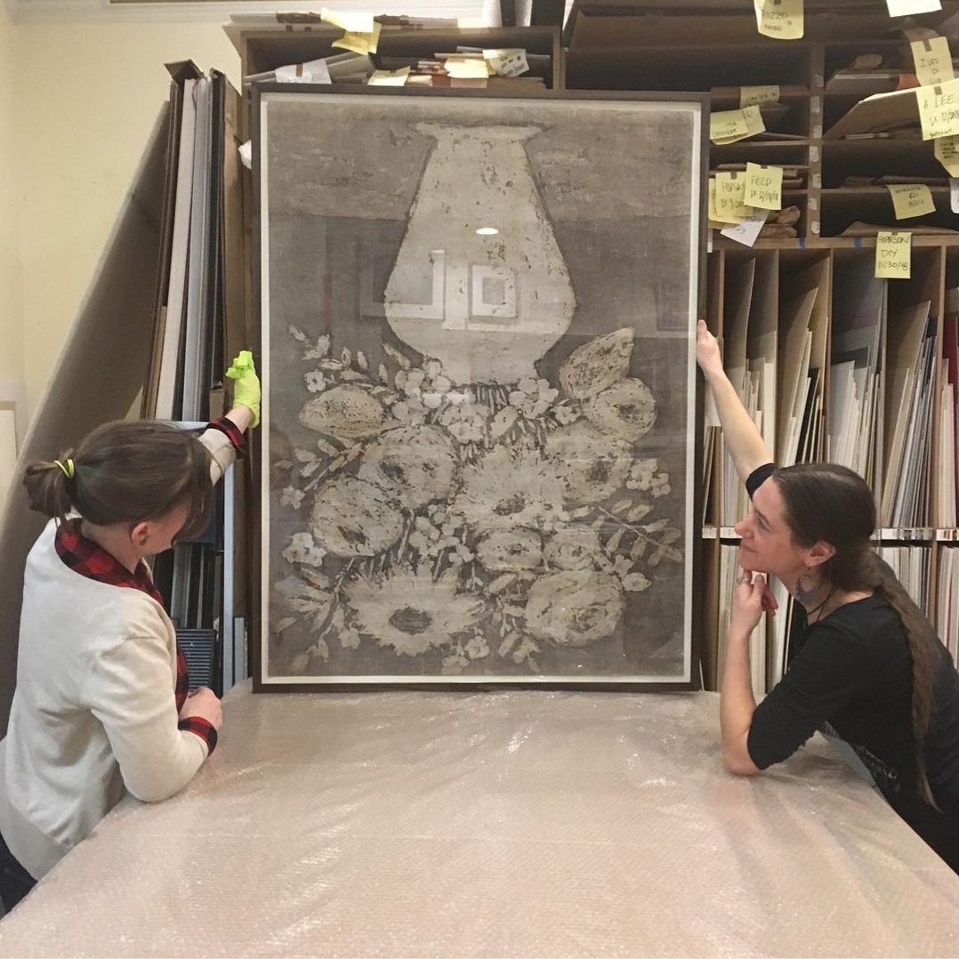 Two women examining an upside-down framed painting of a floral arrangement with a vase, set against a background of stored art supplies and paper in a storage room.