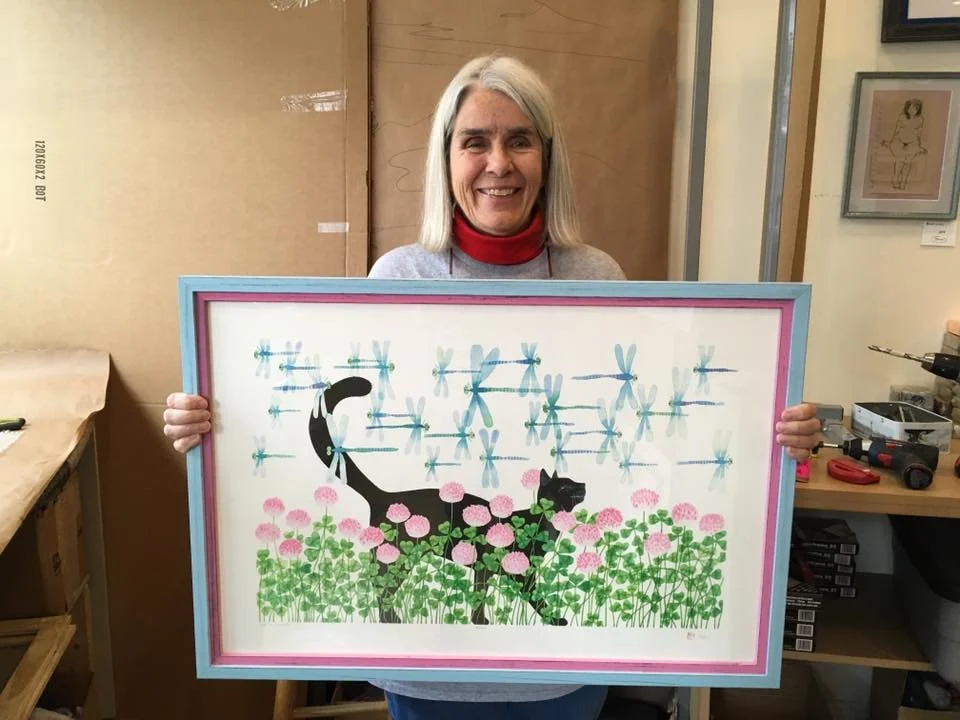 A woman with gray hair smiling and holding a framed art piece depicting a black cat with white whiskers among pink flowers and blue dragonflies.