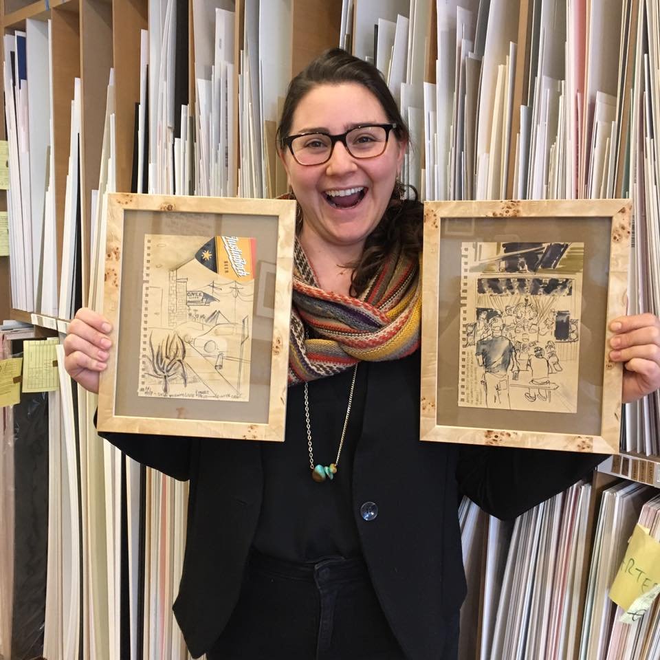 A woman with glasses and dark hair, smiling widely, holds two framed sketches in front of her. She is indoors, surrounded by shelves filled with rolled-up papers or posters.
