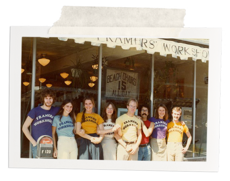 Group of seven young adults standing outside a building with signs that say 'Tramers Workshop' and 'Beach Chairs is All Day', some wearing 'Tramers Workshop' branded T-shirts in yellow, purple, red, blue, and white, sunny weather.