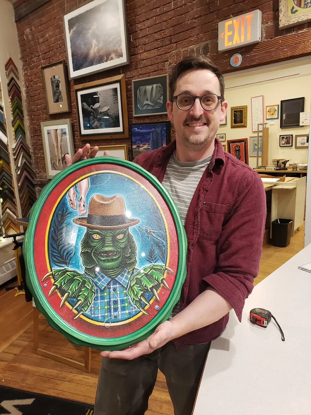 A man is holding an oval art piece featuring a green zombie-like creature with claws, wearing a plaid shirt and hat, set against a colorful background with leaves.