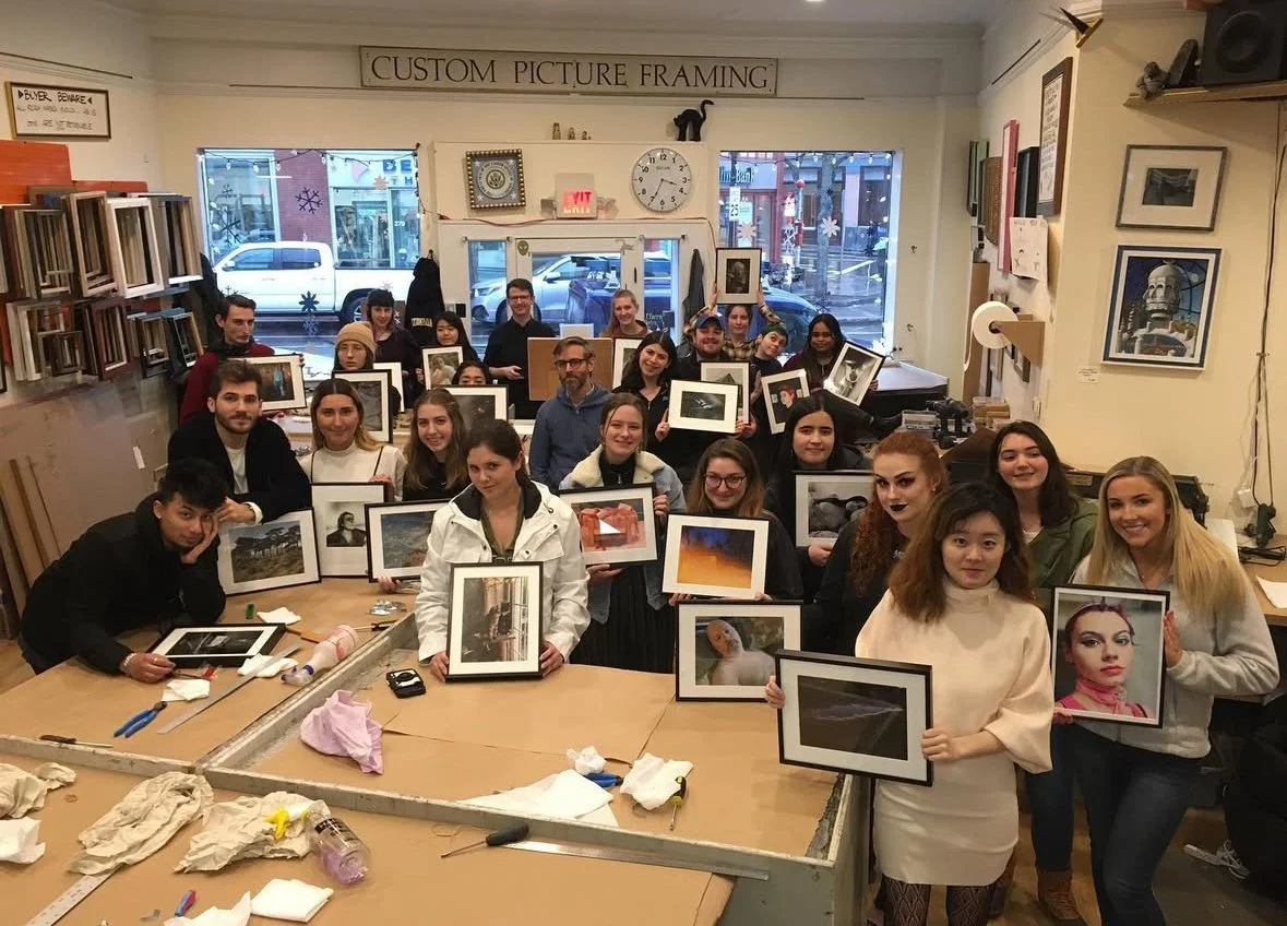 Group of people in an art framing shop holding framed photographs, with art supplies on the table and wall decor