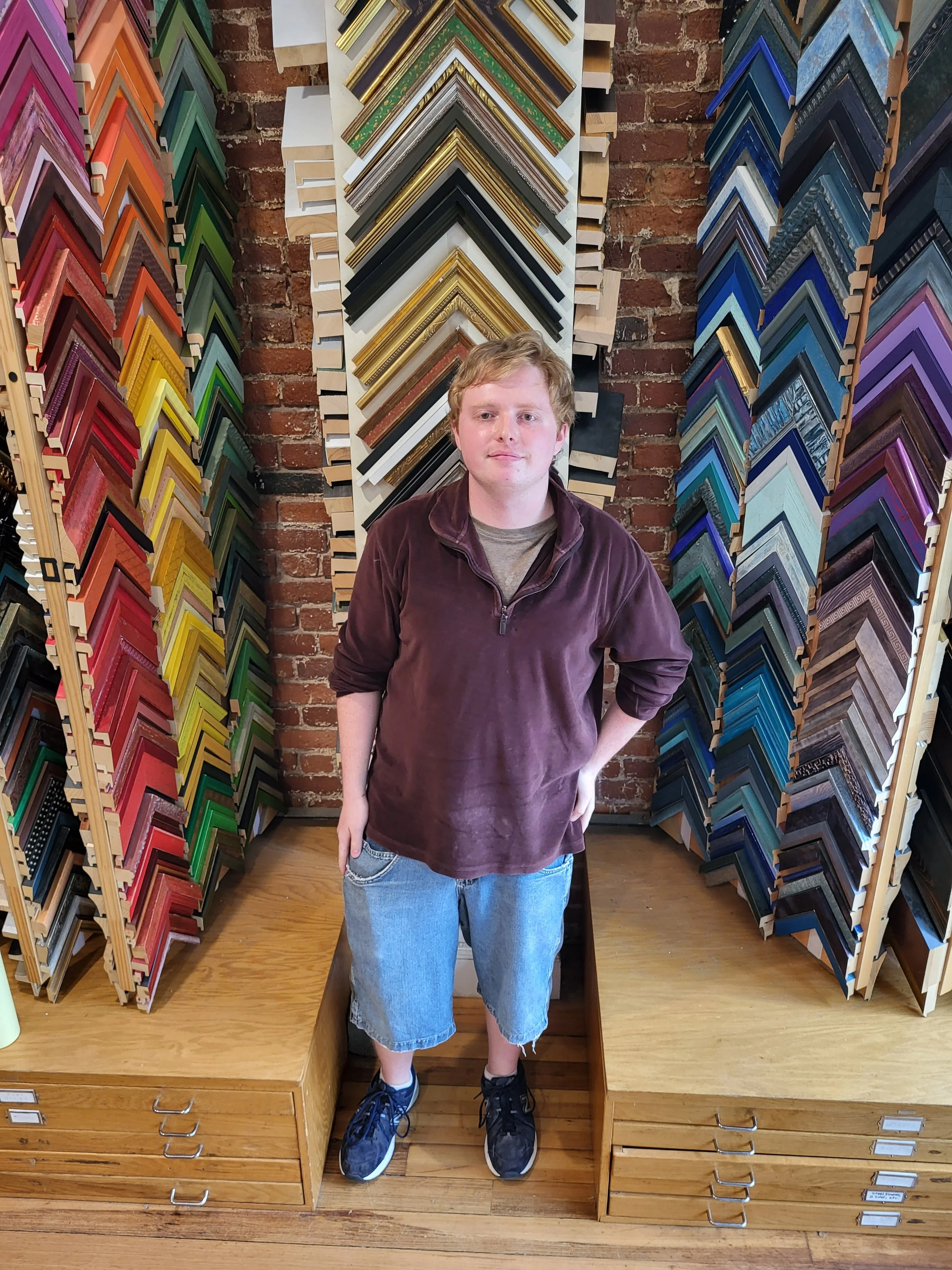 A young man with curly blonde hair wearing a brown velvety jacket, gray T-shirt, and ripped blue jeans, standing inside a frame shop surrounded by various colorful picture frame samples on display.