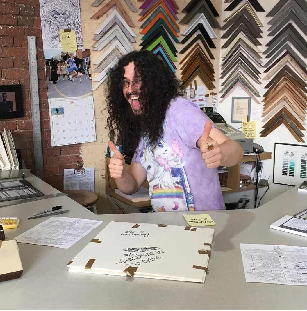 A person with long curly hair and glasses giving two thumbs up while standing behind a desk in an office or art studio with framed artwork, color sample samples, and a calendar on the wall.