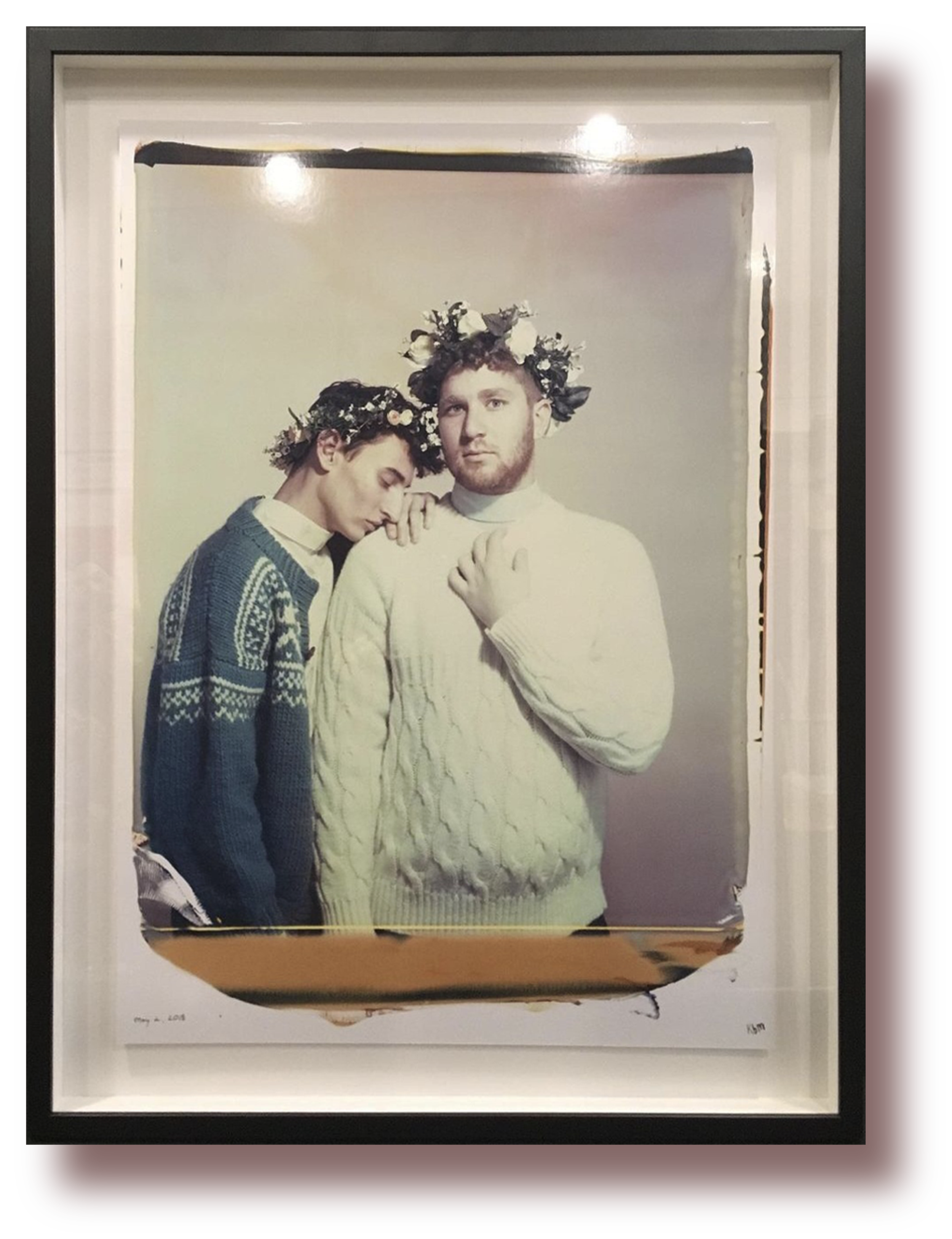 Two young men wearing flower crowns, one in a gray sweater with a patterned design and the other in a white cable-knit sweater, standing close together against a plain light background.