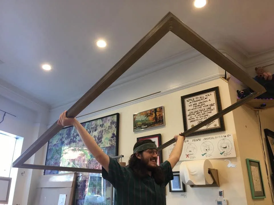 A man in a black shirt and headband holds a large picture frame above his head, as if he is lifting it. He is smiling and standing in a room with various framed pictures and signs on the wall.