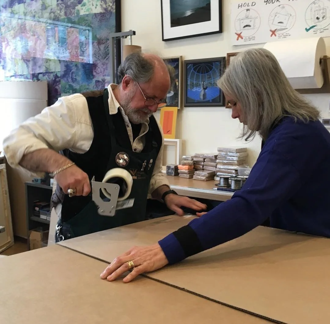 A man and a woman are working together on a craft project at a table. The man is using a handheld tape dispenser, and the woman is holding the cardboard with her hands. The background shows shelves with stacks of materials and a wall with instruction
