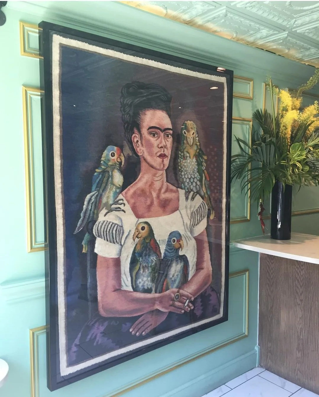 Framed artwork of a woman with dark hair styled in an updo, surrounded by colorful parrots, displayed on a green wall with gold trim.