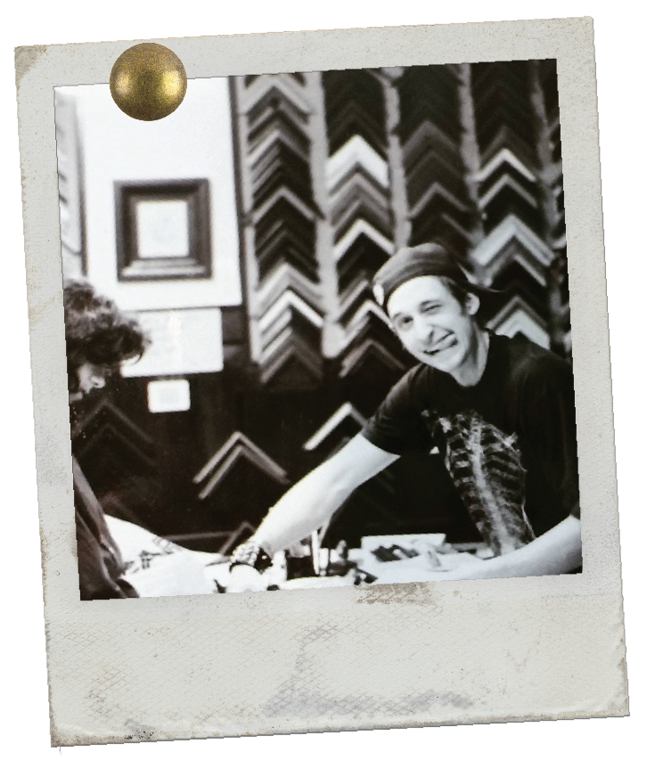 A black and white photo of a young man with a hat and a graphic t-shirt smiling and extending his hand, inside a frame with a gold ball pin and frame samples in the background.