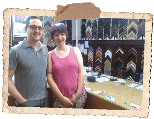 A man and woman standing inside a frame shop, smiling, with framing samples on the wall behind them.
