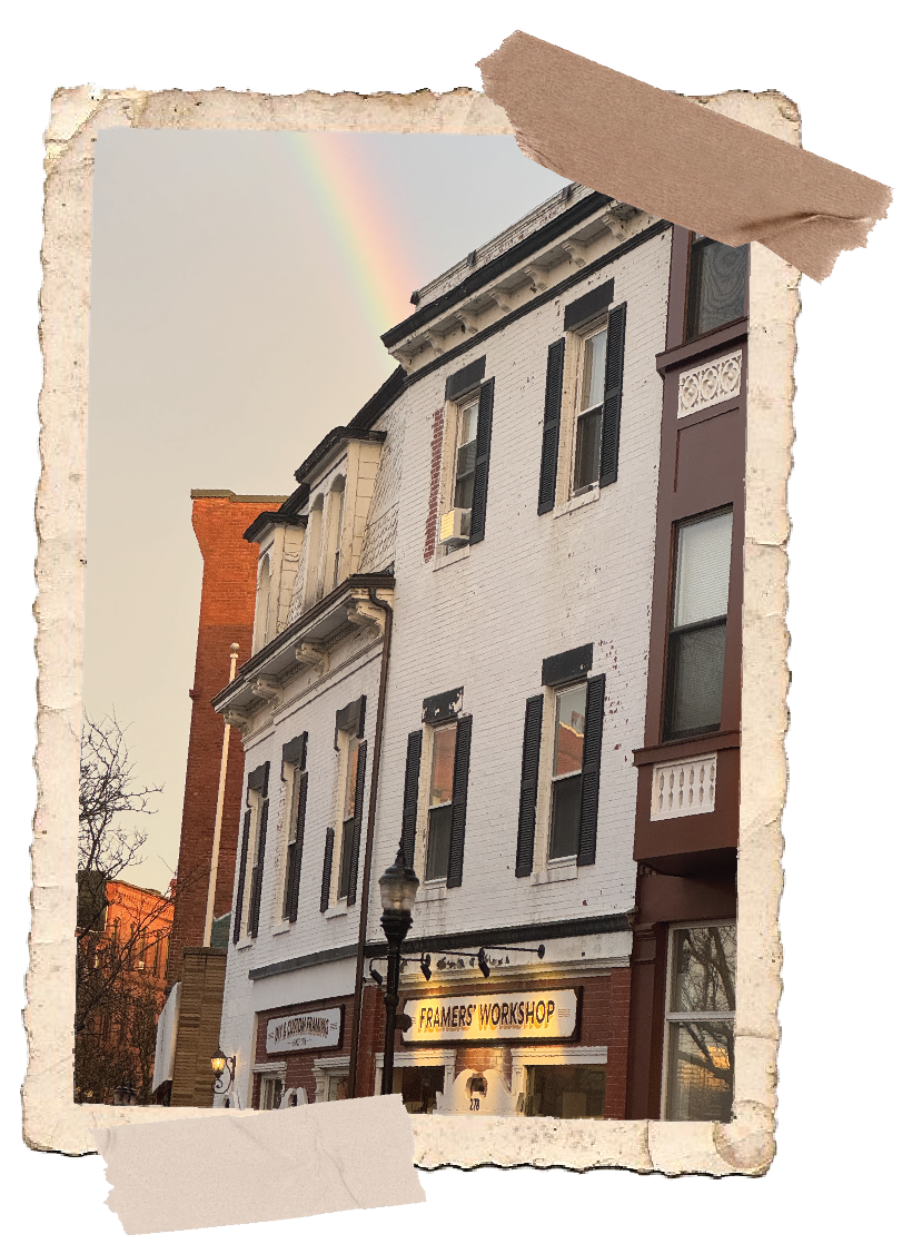 A three-story brick building with a white facade, black window shutters, and a shop on the ground floor with a lit sign that reads "Framers' Workshop." There is a lamppost in front of the building, and a rainbow in the cloudy sky above.