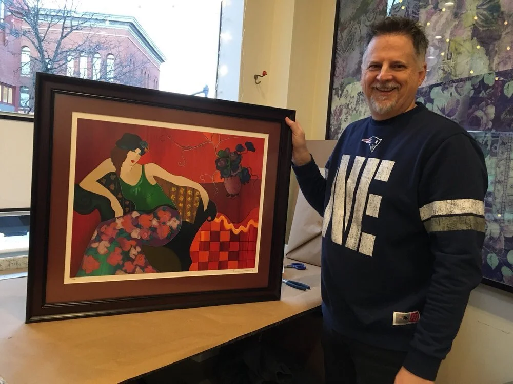 A man standing indoors near a large framed painting depicting a woman sitting on a sofa with a flower on her lap and potted plants nearby.
