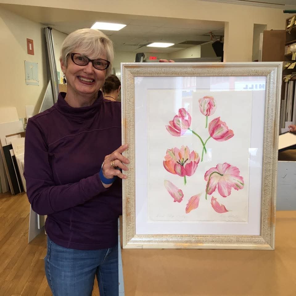 A woman with short gray hair, glasses, and a purple top smiling and holding a framed watercolor painting of pink tulips in a bright room.