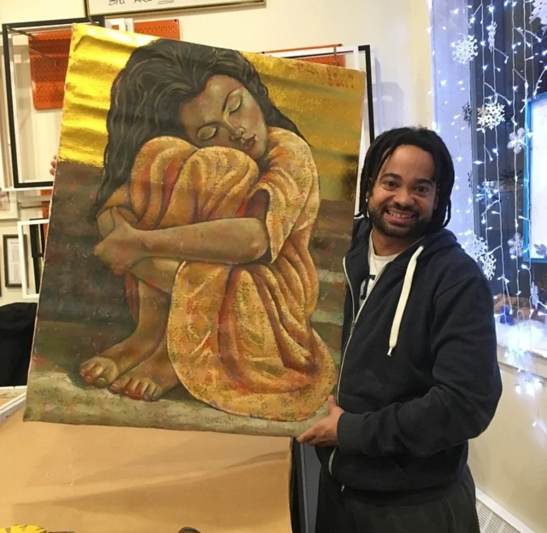 A man with dreadlocks smiling and standing next to a large painting of a curled-up woman with long dark hair, embracing her knees with her eyes closed. The setting appears to be an art gallery or studio with Christmas decorations and framed artwork o
