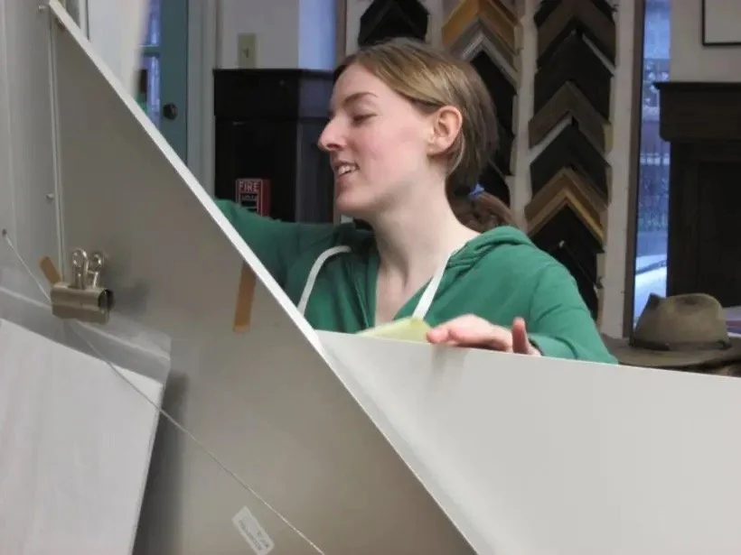 Young woman with brown hair in a ponytail, wearing a green hoodie, is shopping for framing materials in an art store.