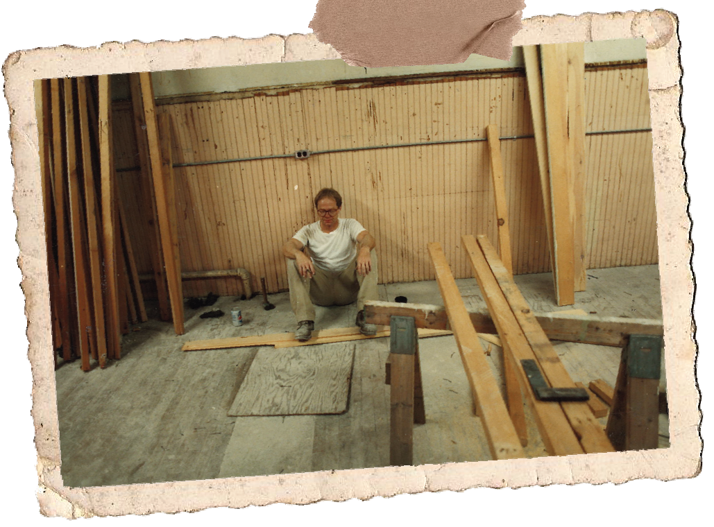 An old image of the owner sitting cross-legged on the floor of the construction site of the workshop, surrounded by wooden planks and construction materials.