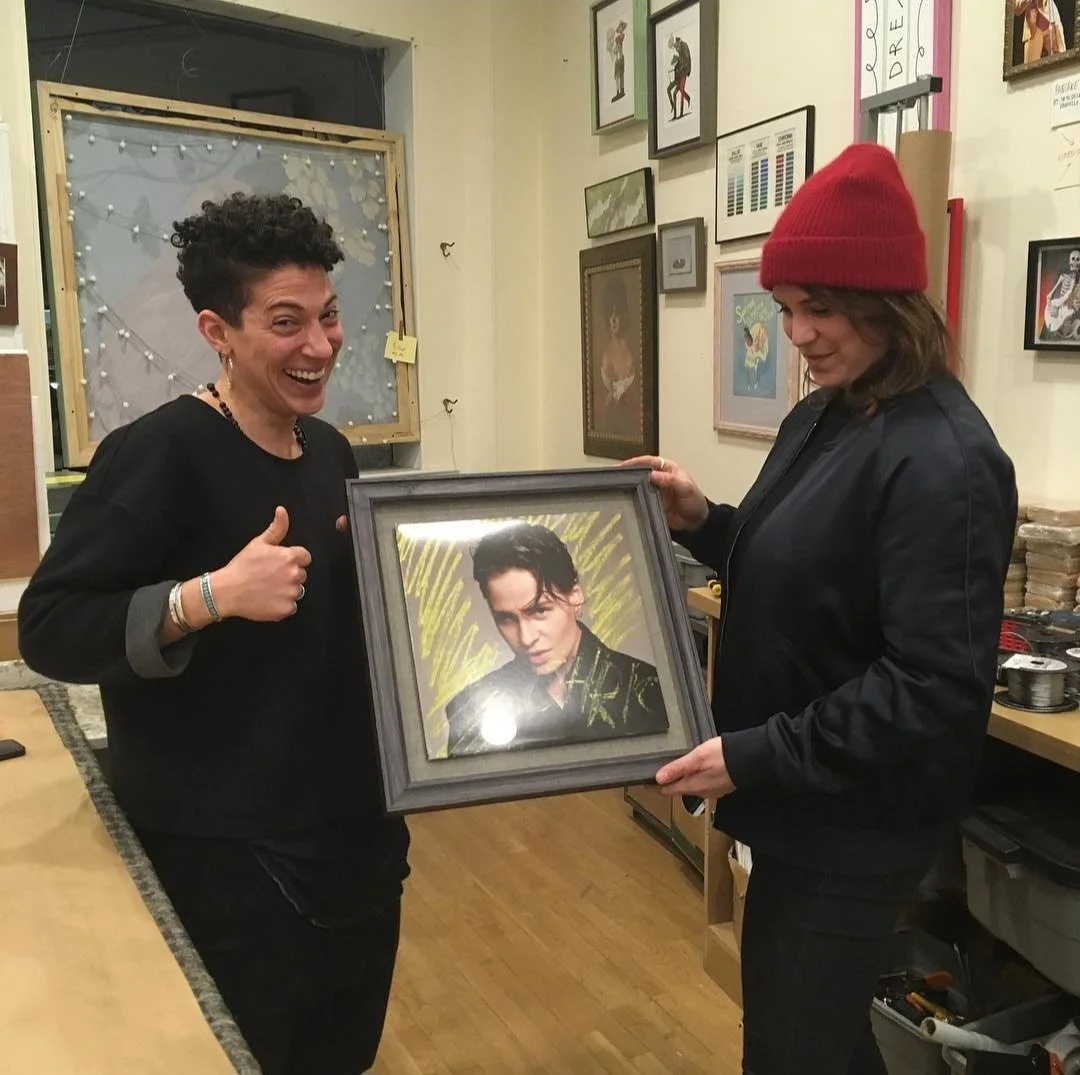 Two women in a room holding a framed portrait of Michael Jackson. One woman is smiling and giving a thumbs up, while the other woman is looking at the portrait. The room has various artwork and frames on the walls.